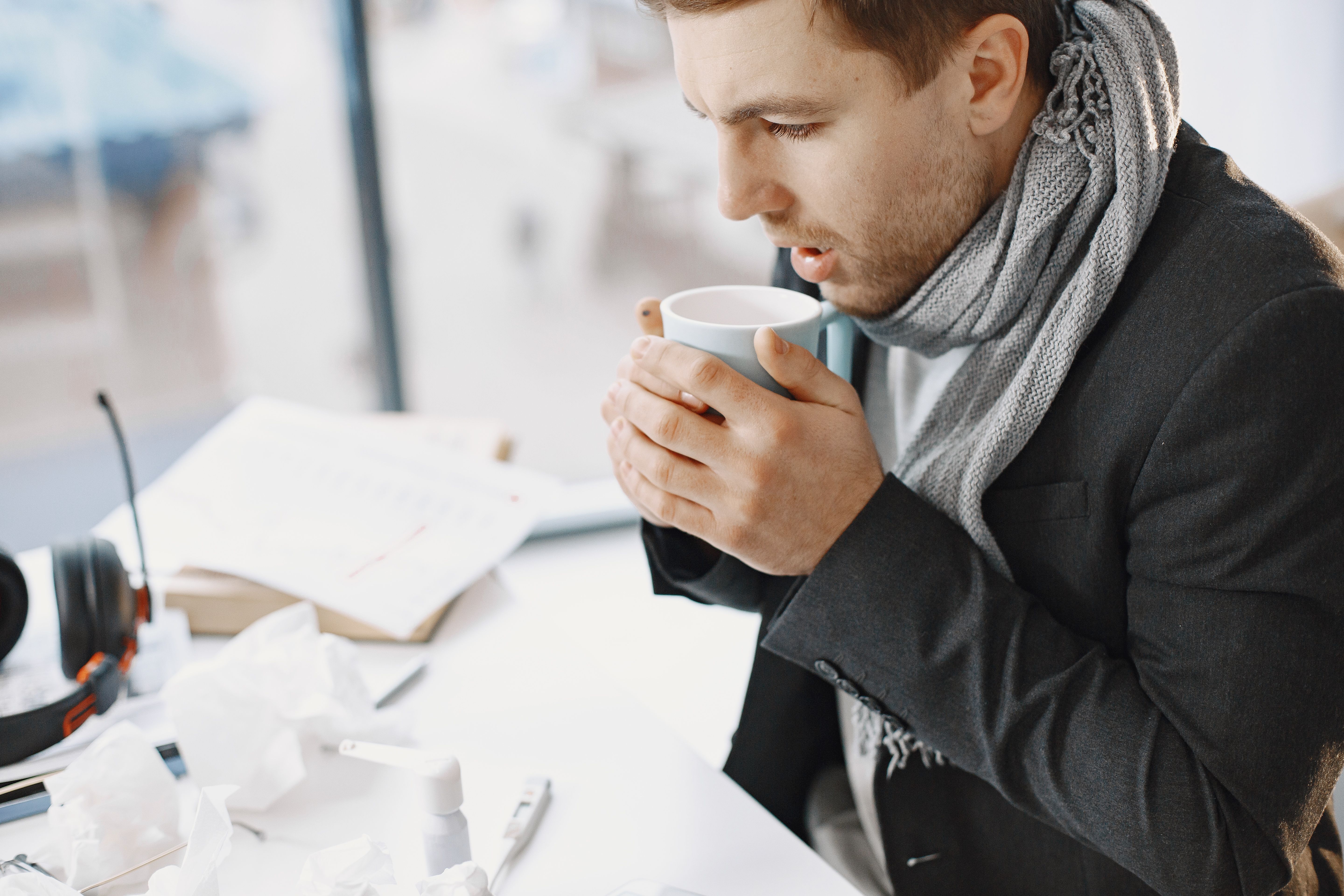 A man wearing a grey scarf and jacket sitting at a desk holding a warm mug while surrounded by tissue paper and medicine bottles.