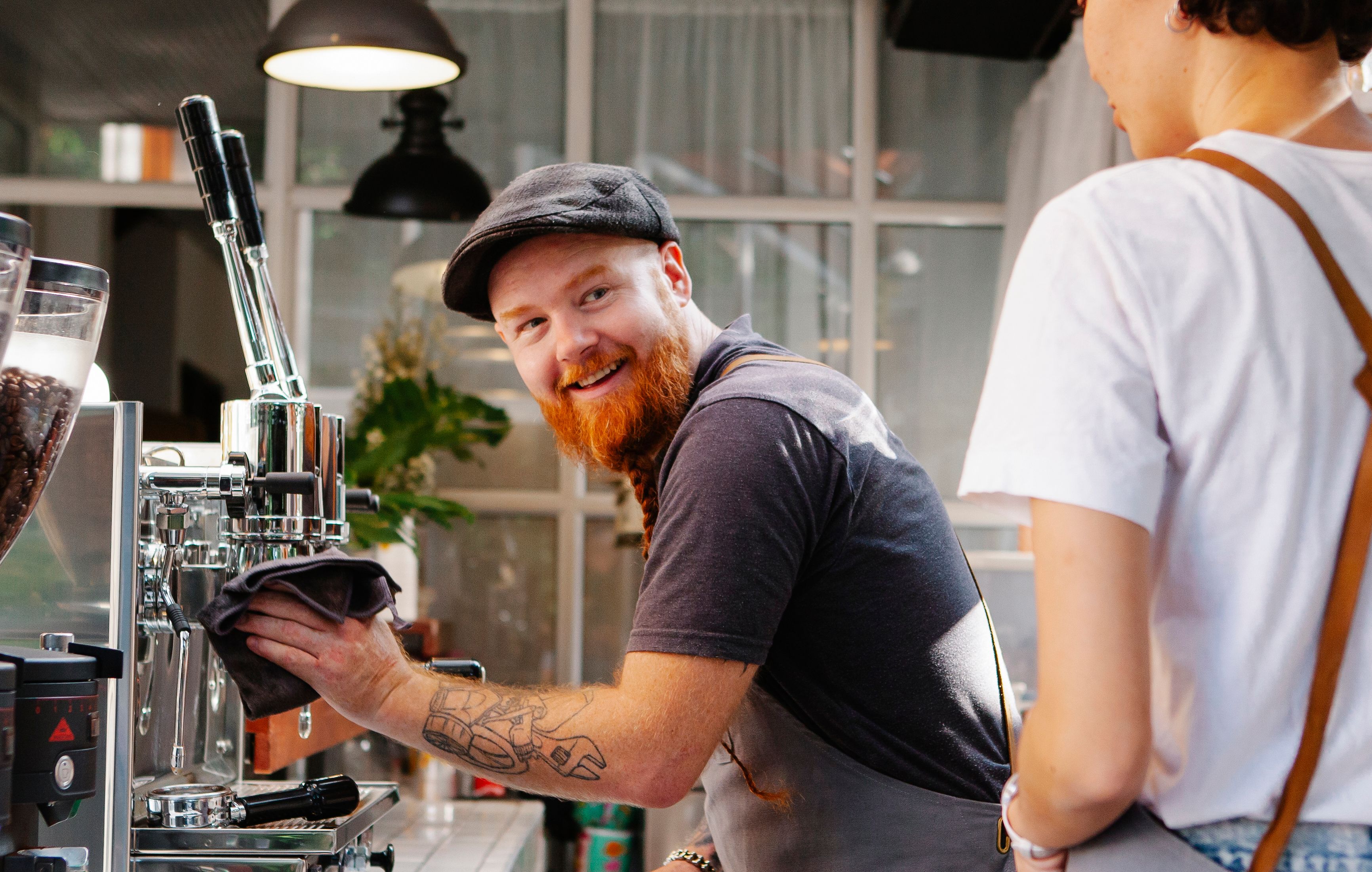 A smiling red-bearded barista wearing a flat cap cleaning a commercial espresso machine with a cloth while interacting with a coworker.