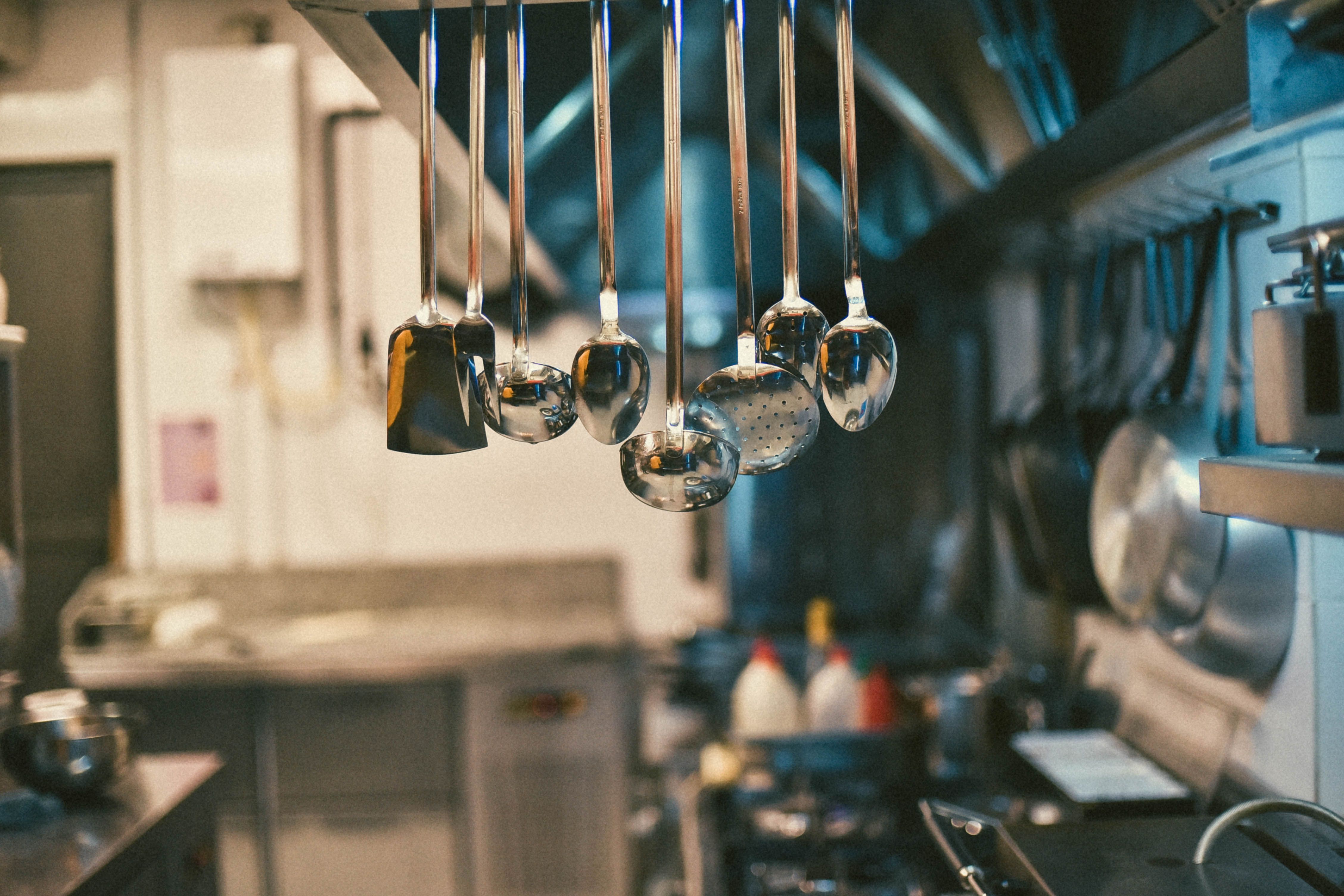 Stainless steel cooking utensils including ladles and spatulas hanging in a row inside a commercial kitchen.