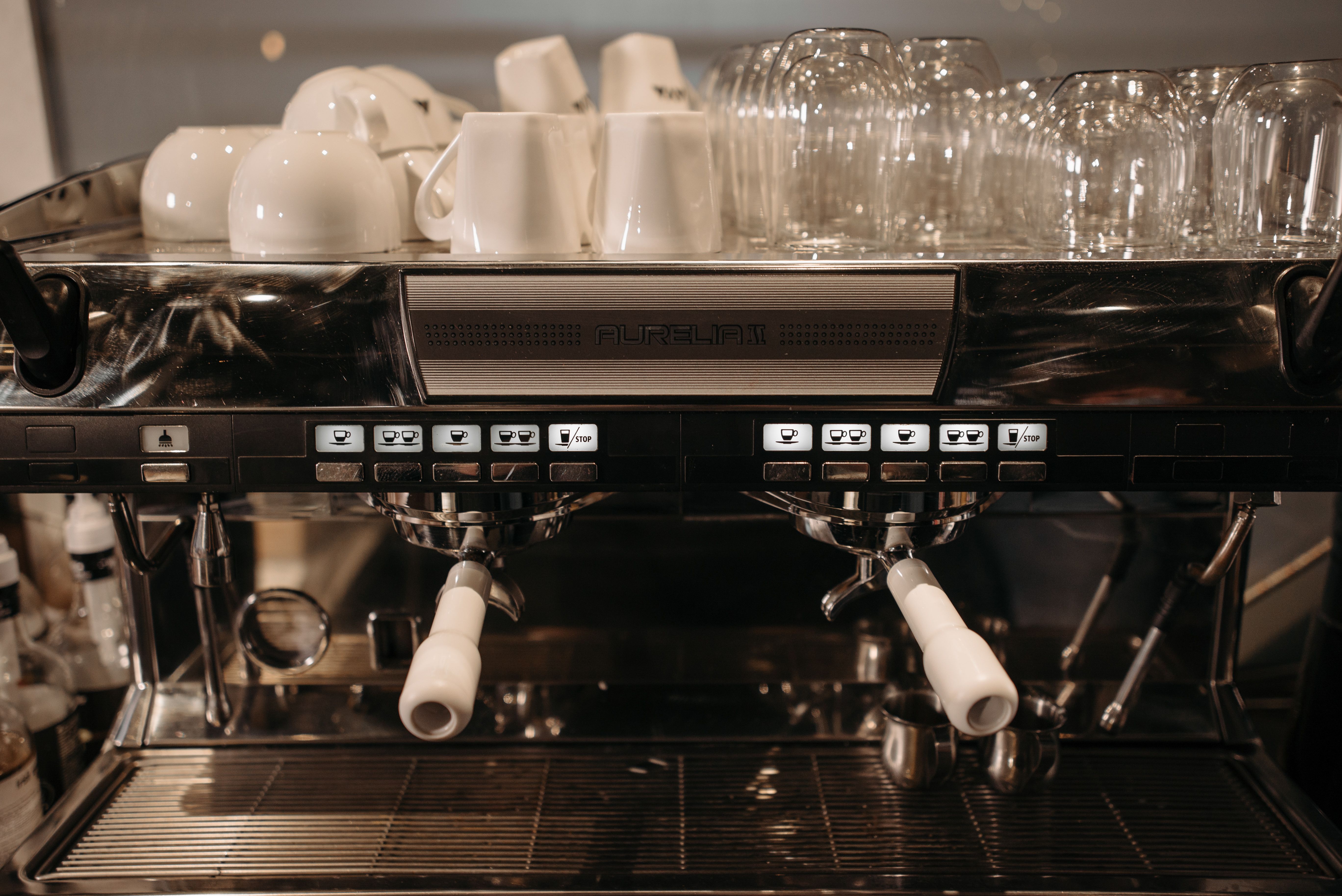 Close-up of a commercial espresso machine with white porcelain cups stacked on top and glass cups stored beside them.