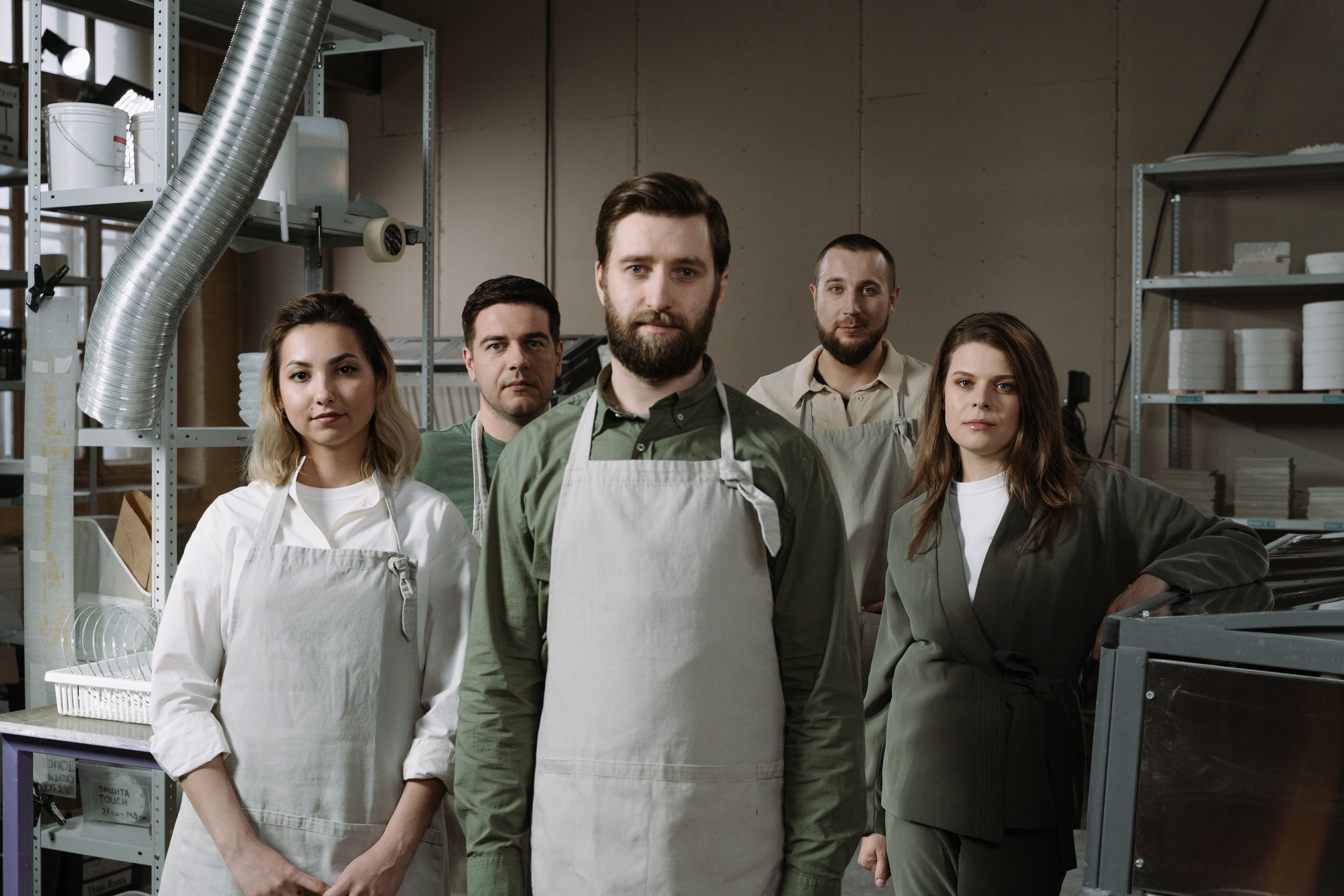 Group portrait of five kitchen team members wearing aprons standing together in a workshop or studio setting.