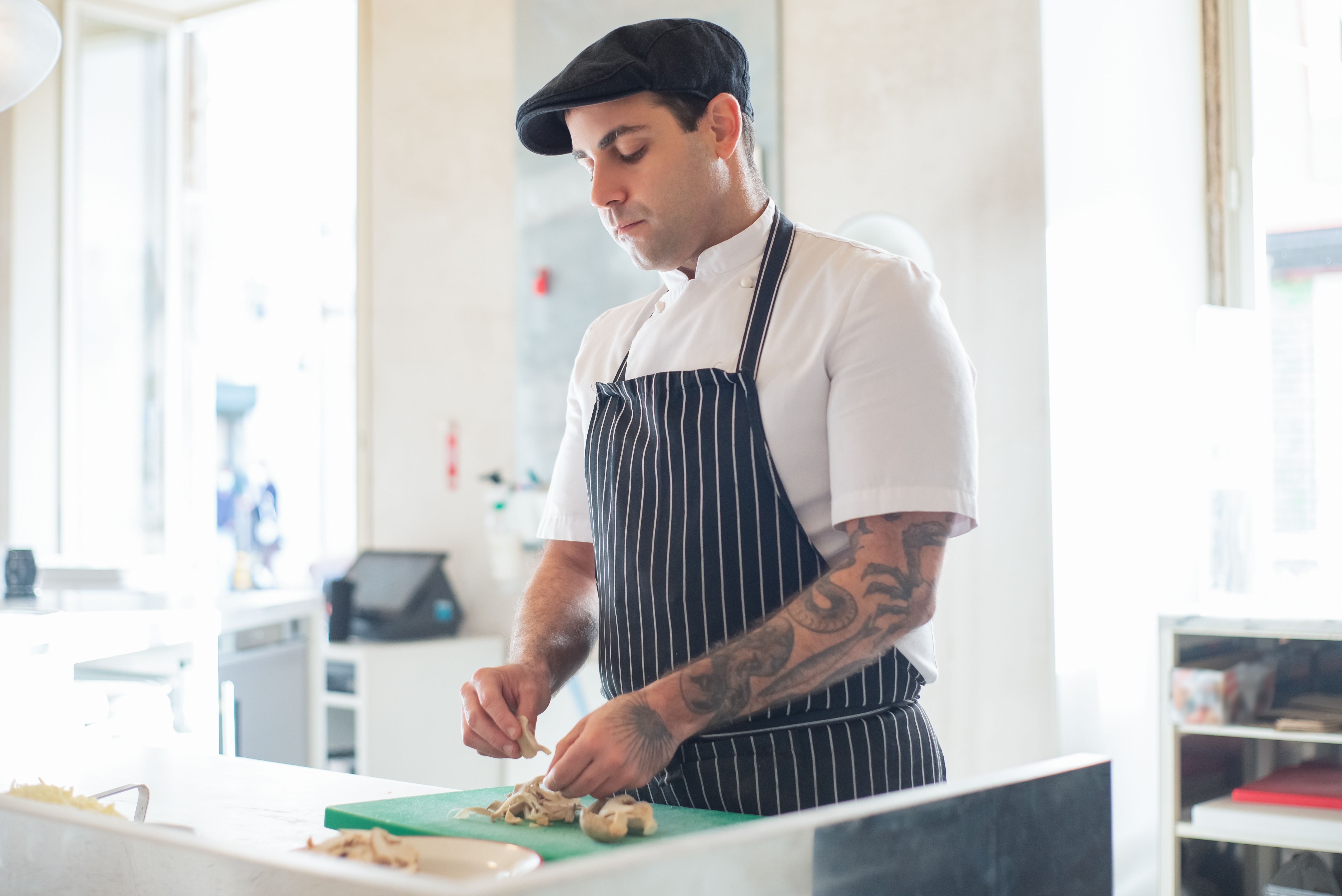 Chef wearing a flat cap and striped apron meticulously prepping ingredients on a green cutting board at a kitchen counter.