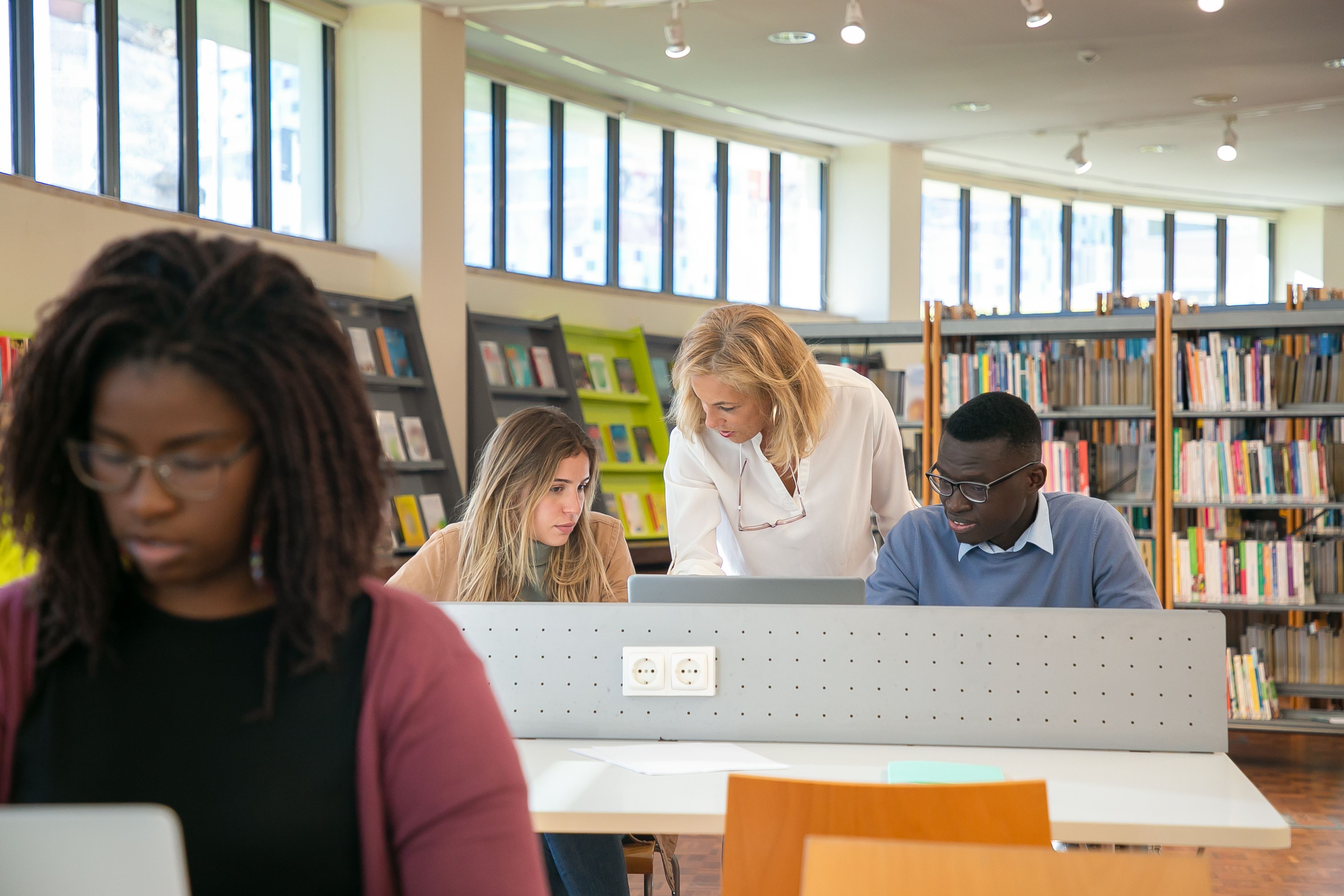 Students working at study tables in a brightly lit library while a teacher assists a student with a laptop.