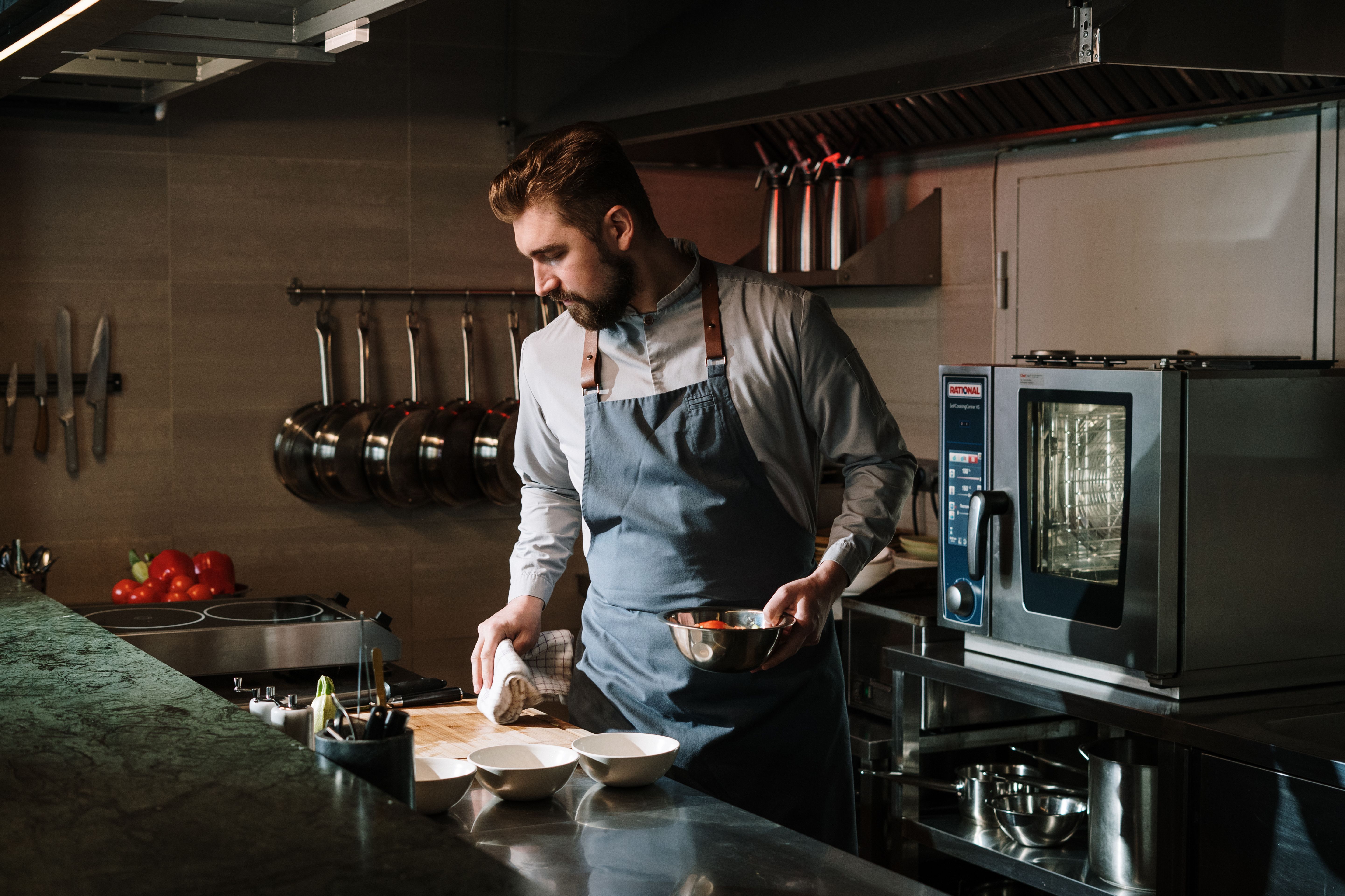 Male chef in an apron wiping down a preparation counter next to a commercial combi oven in a modern kitchen.