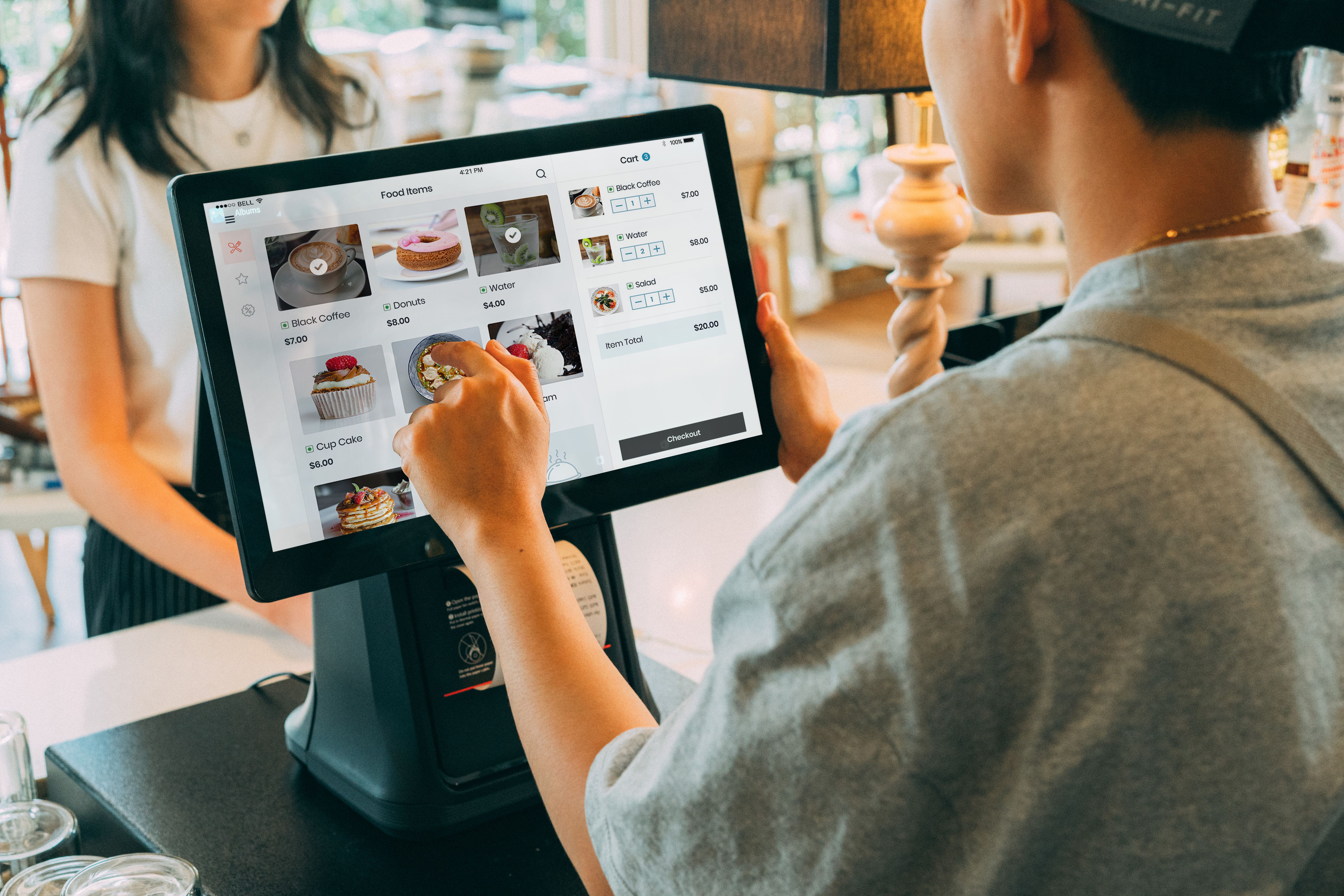 Over-the-shoulder view of a cashier using a digital point-of-sale tablet to select menu items at a store counter.