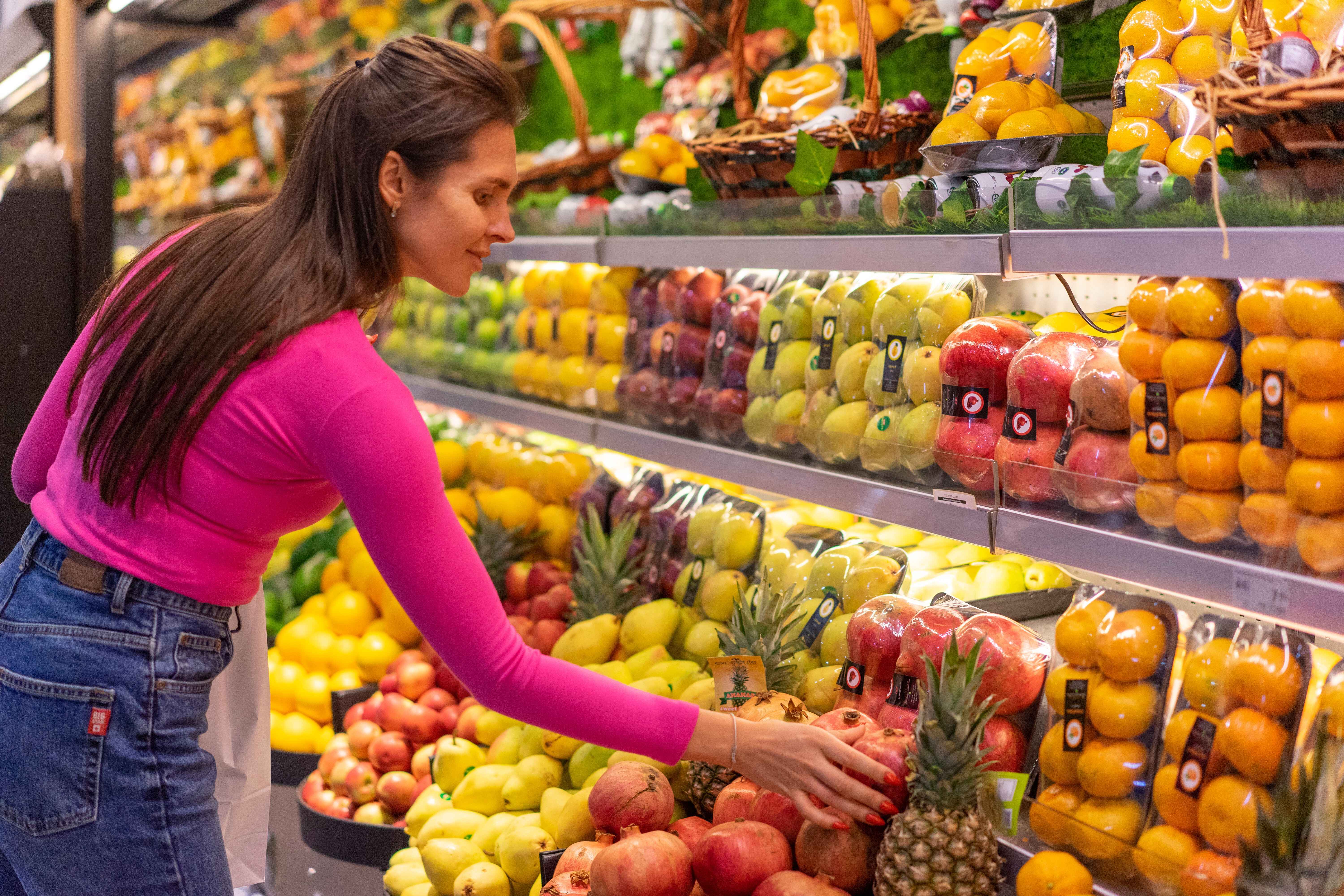 Woman in a bright pink top selecting fresh pomegranates in a supermarket produce aisle.