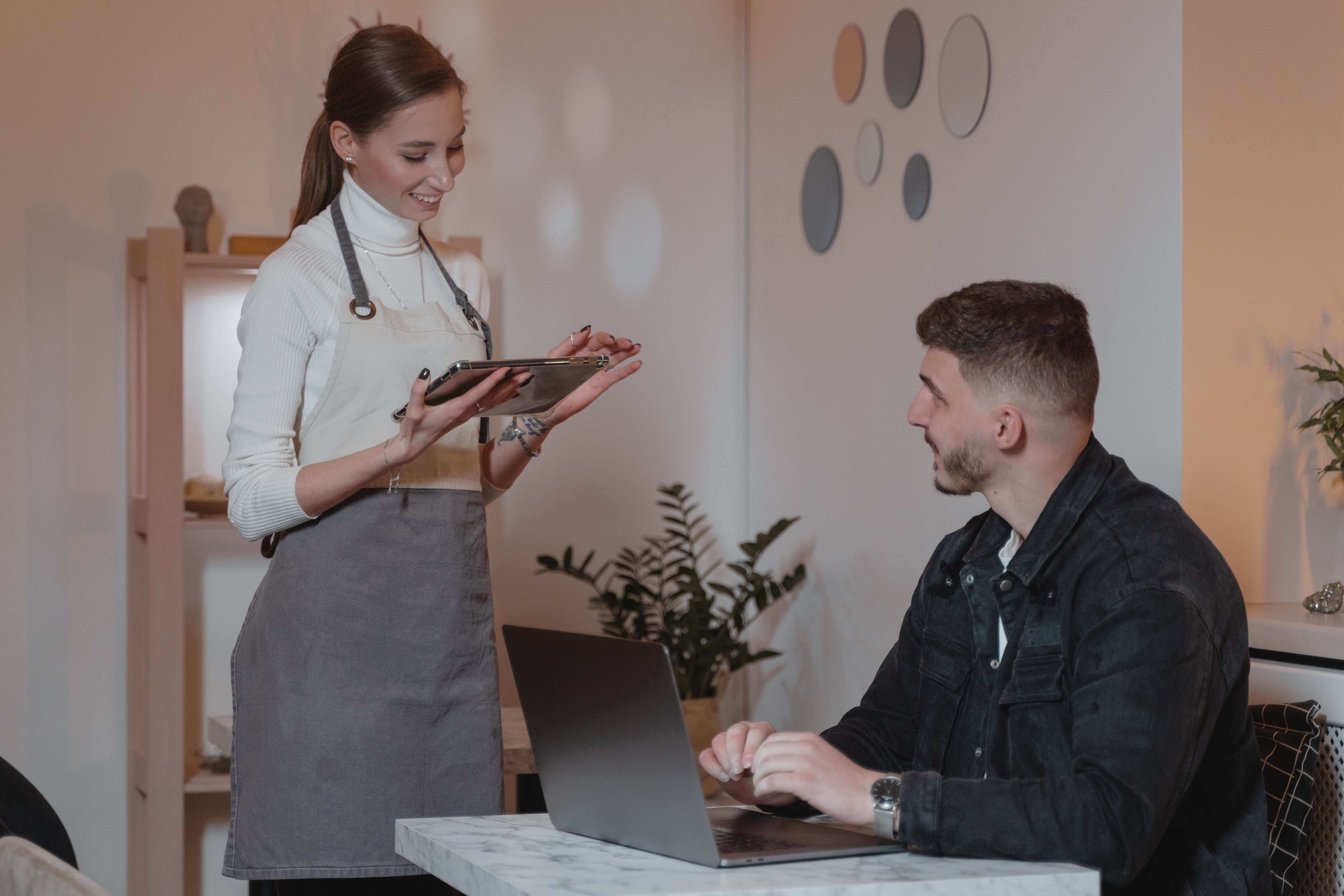 Waitress in an apron using a digital tablet while taking an order from a seated customer with a laptop.