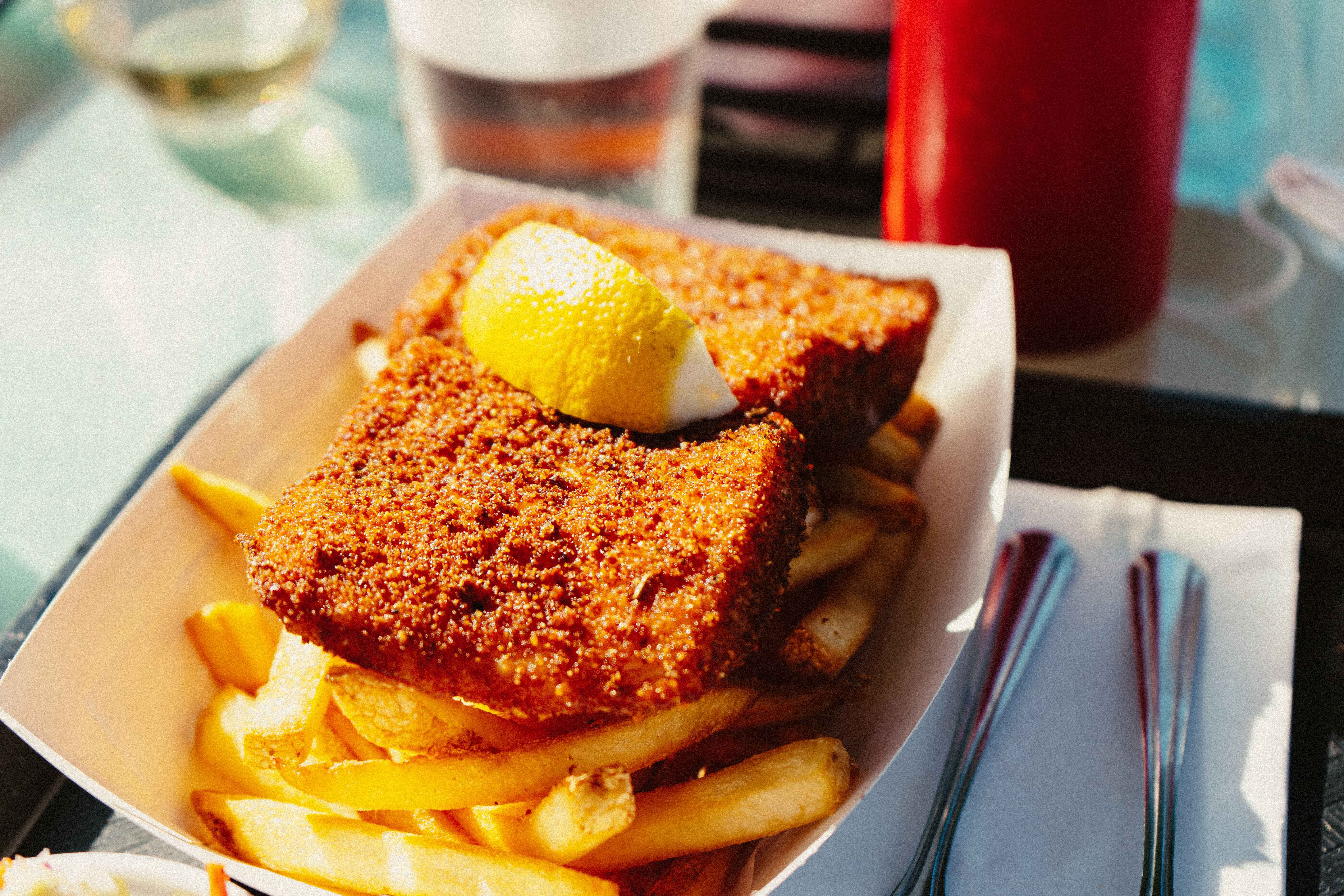 Breaded vegan fish substitute topped with a lemon wedge resting over a bed of French fries in a paper tray.