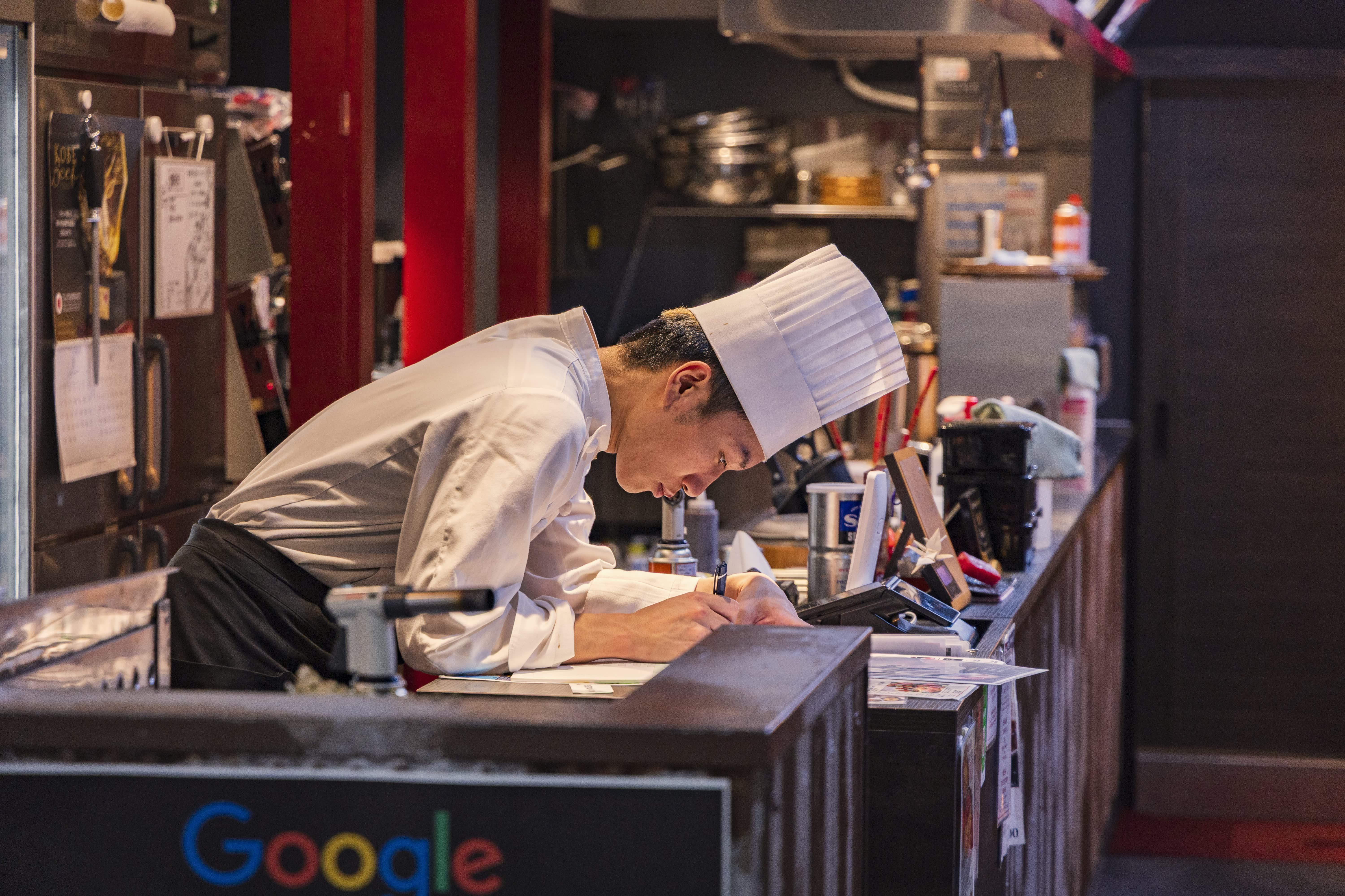 A chef wearing a tall chef hat writing notes on paper over a service counter inside a restaurant kitchen.