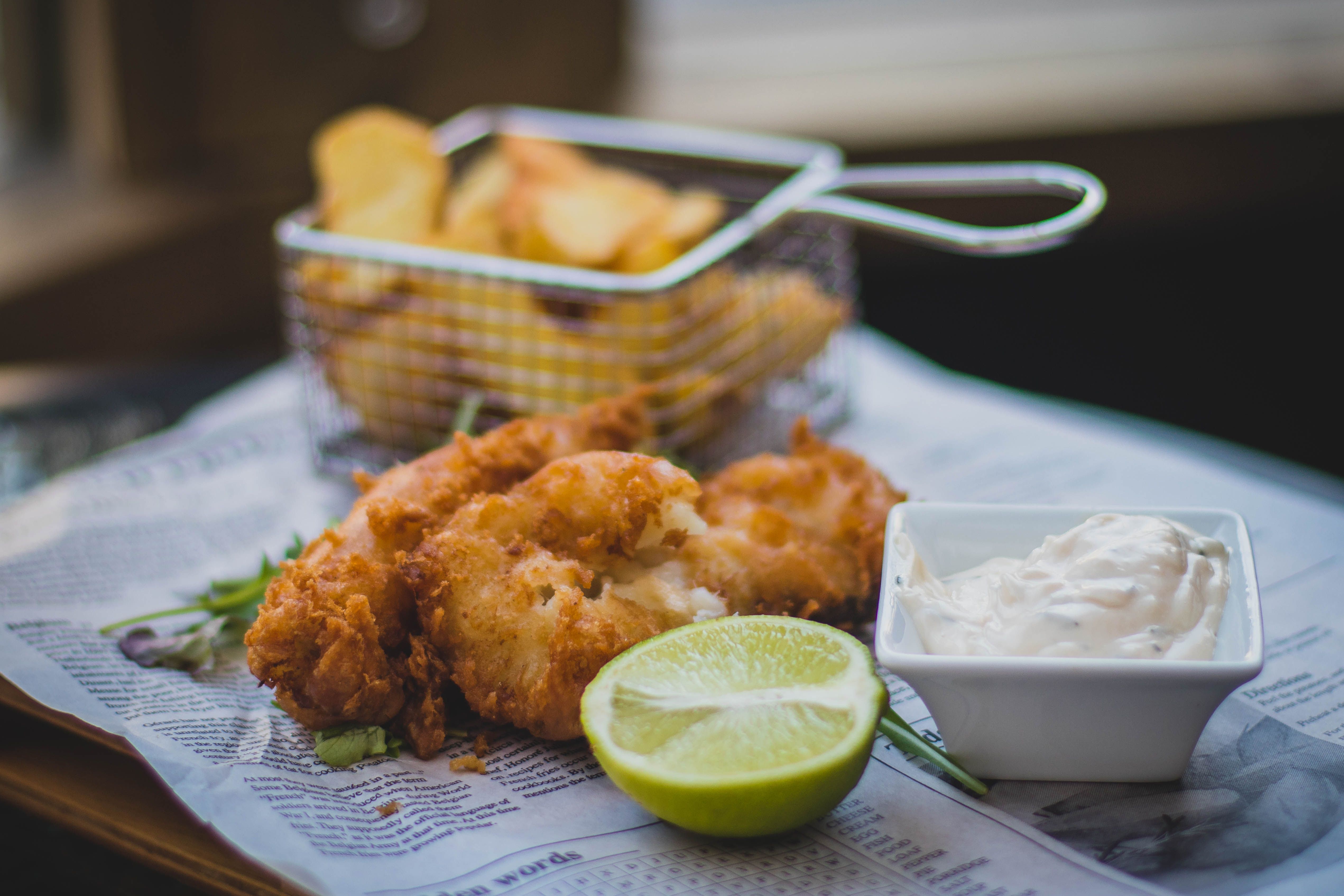 Golden-battered fish fillets served with a lime wedge, dipping sauce, and french fries in a wire basket over newsprint paper.