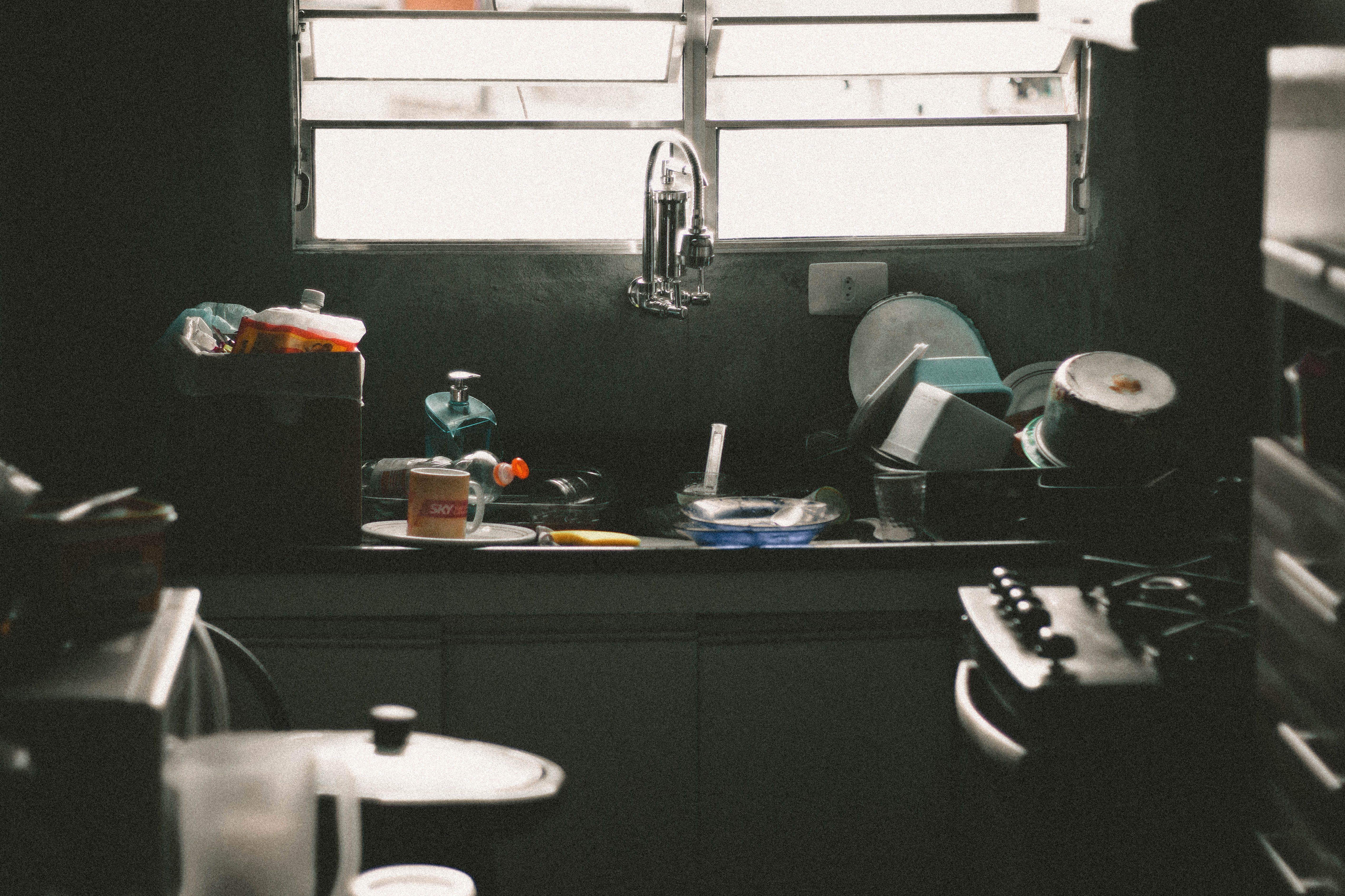 Cluttered residential kitchen sink area filled with unwashed dishes and utensils below a window.