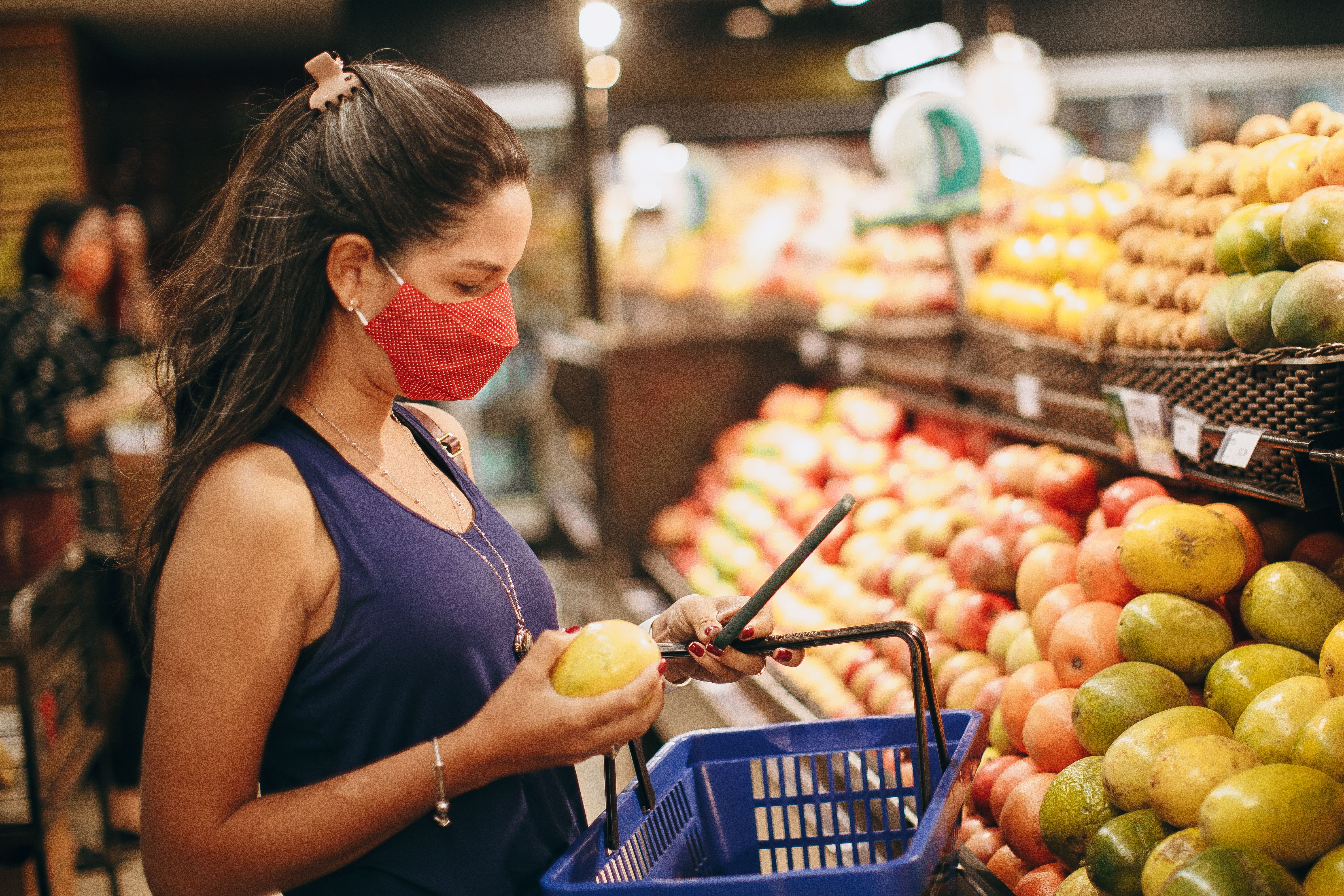 Woman wearing a red protective face mask holding a lemon and checking her phone in a supermarket produce aisle.