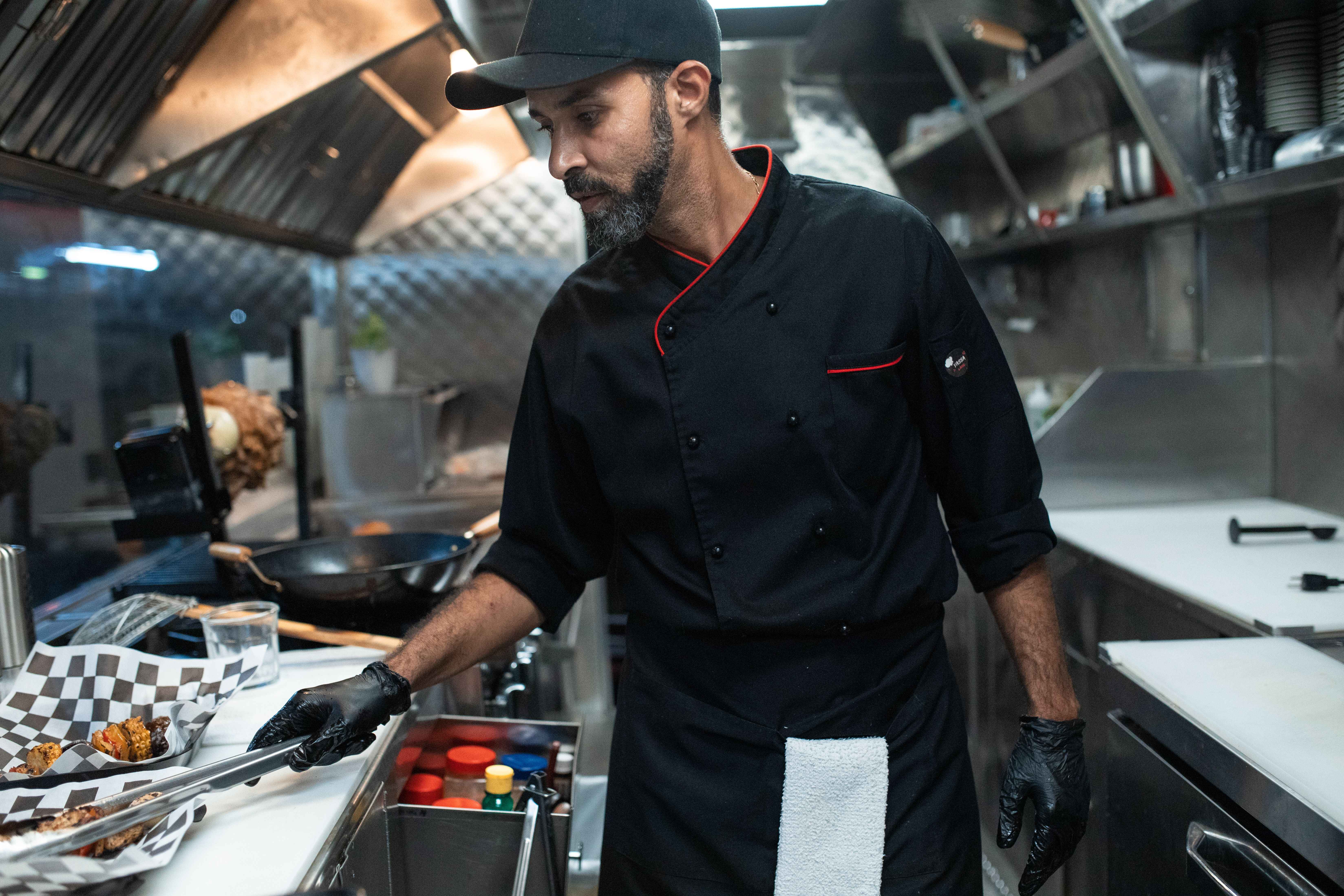 Male chef wearing a black chef jacket, cap, and black gloves using tongs to serve food in a commercial kitchen.