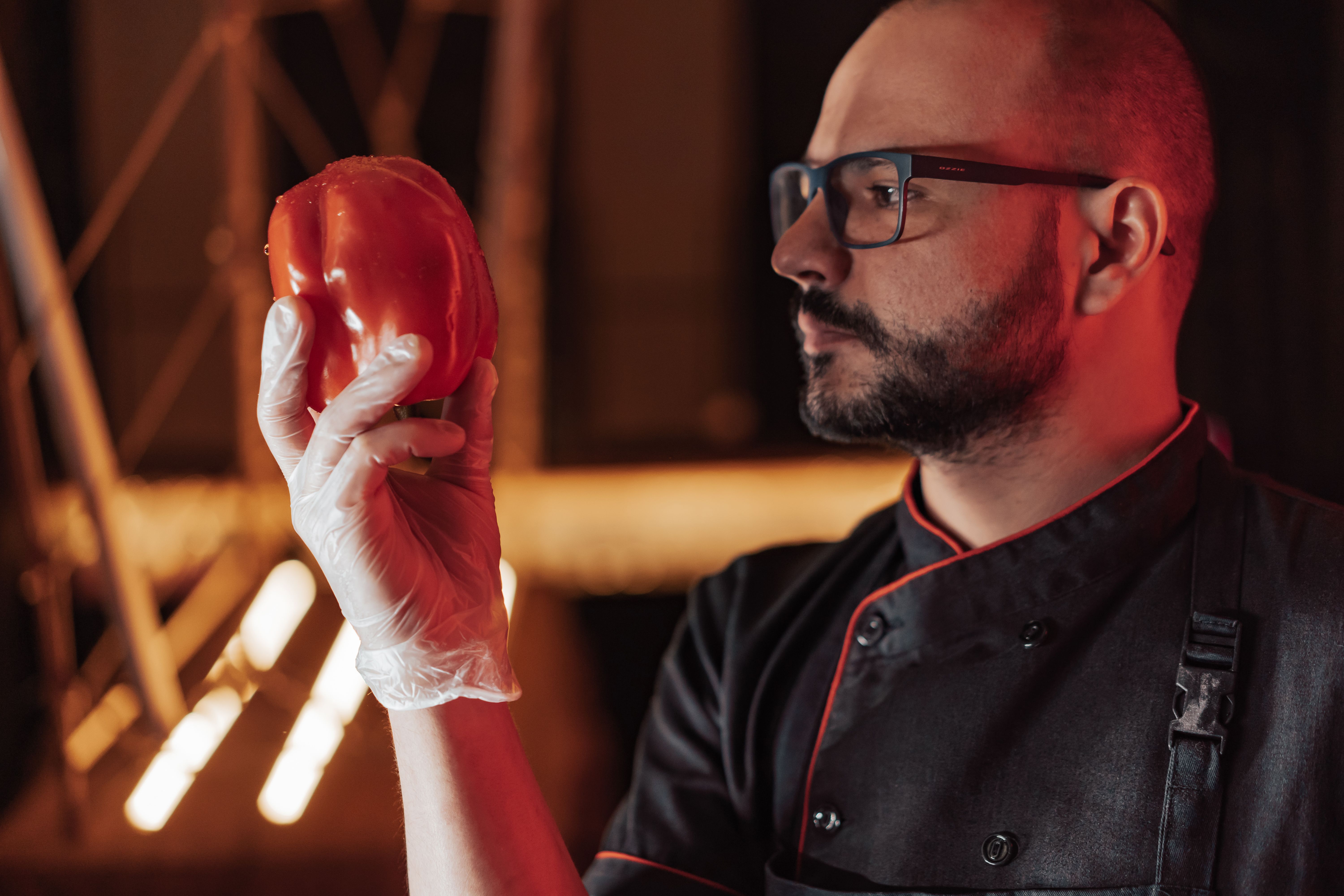 Male chef wearing glasses, a black jacket, and clear disposable gloves inspecting a red bell pepper.