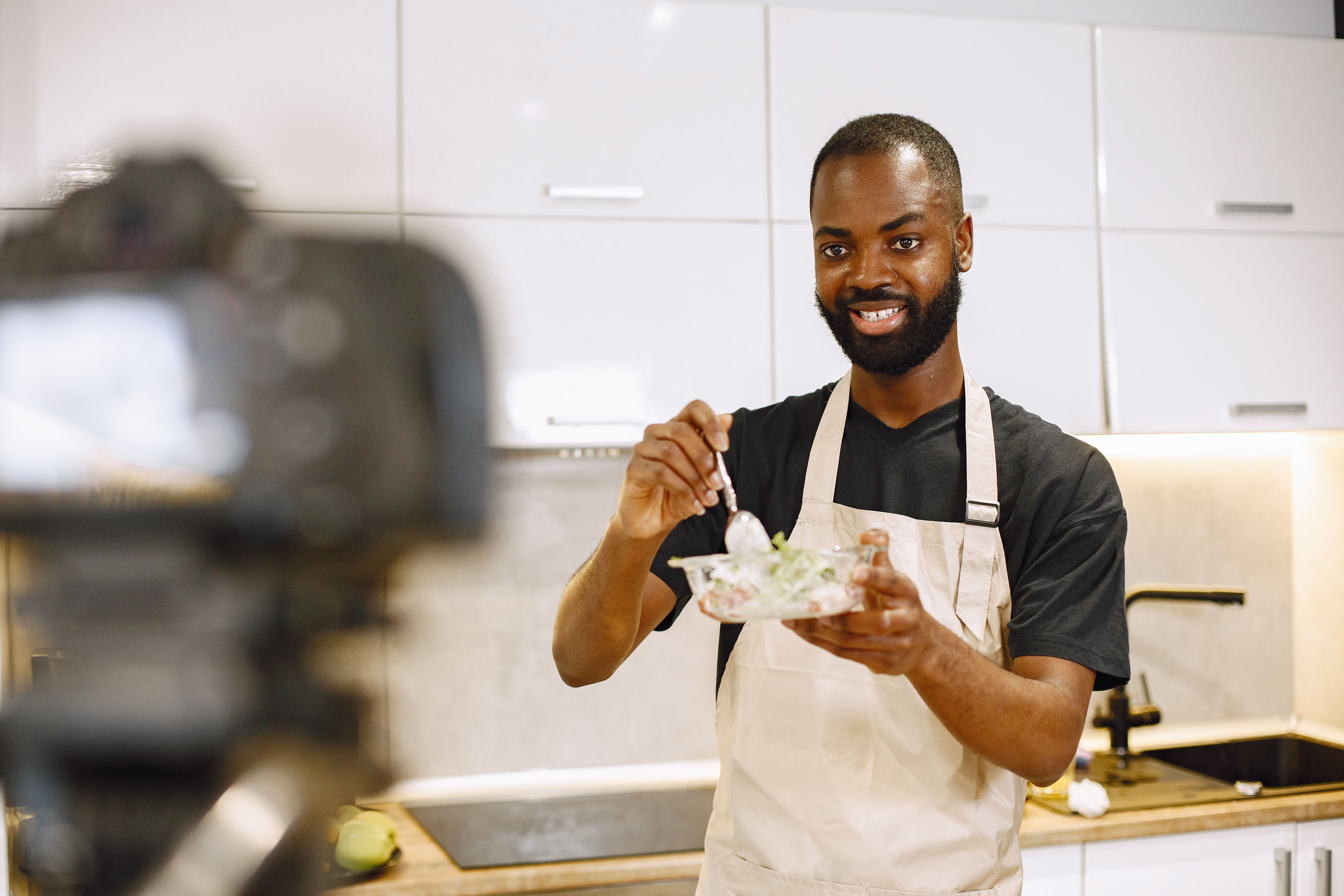 Man in a light apron holding a clear bowl of salad and a spoon while filming a cooking video in front of a camera.