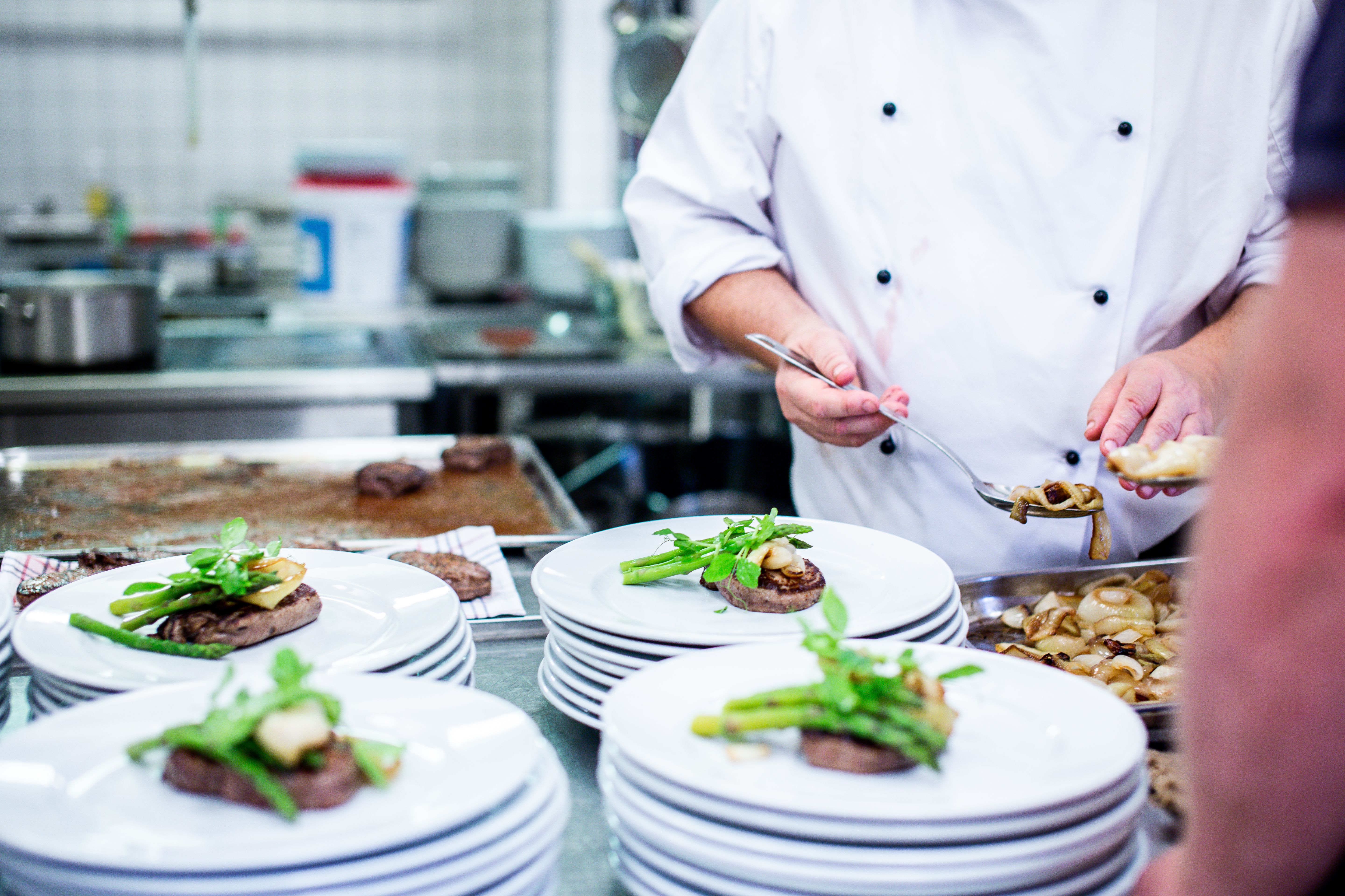 Chef in a white double-breasted coat using a spoon to plate food onto steak dishes topped with asparagus.