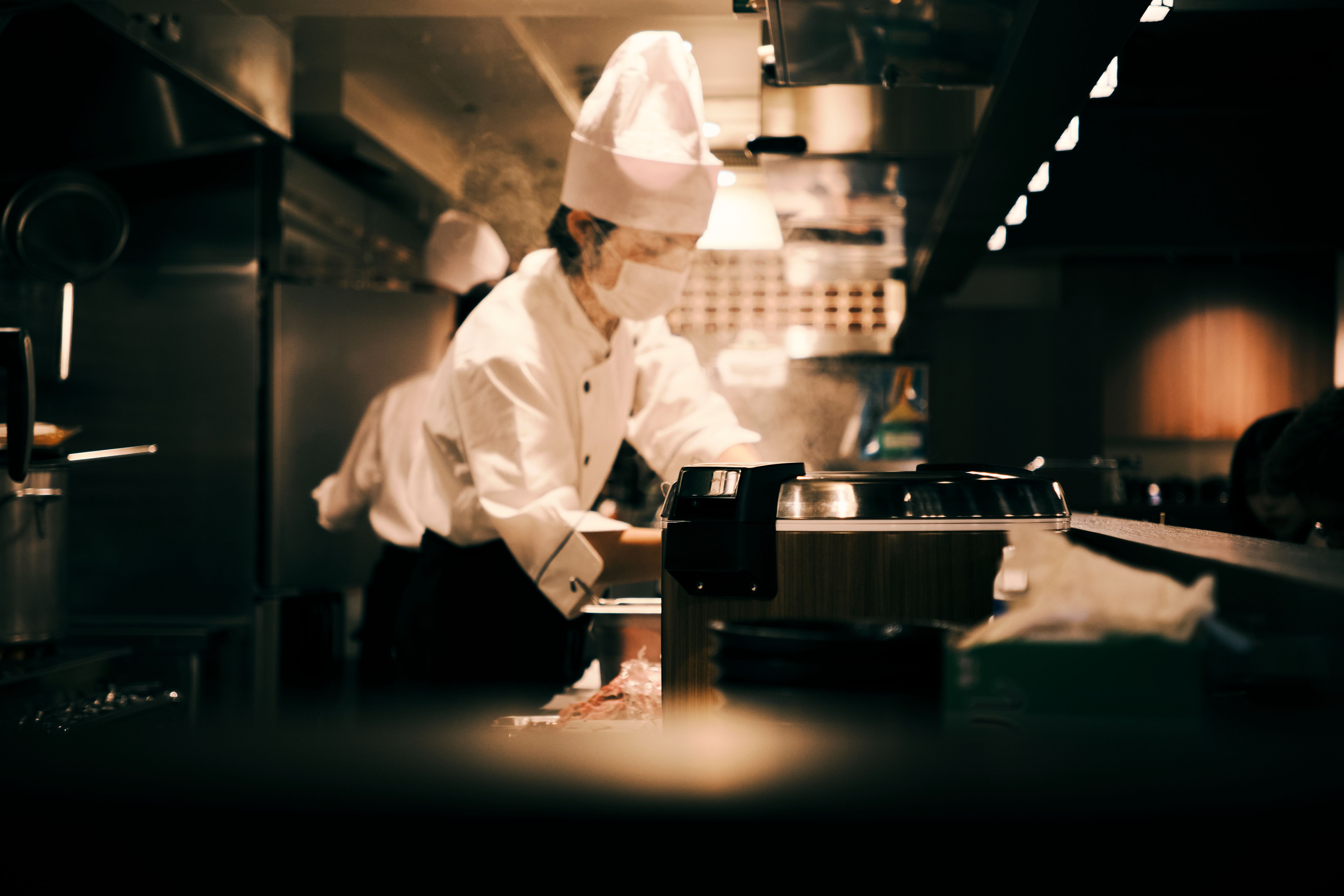 Chef wearing a tall chef hat and face mask preparing food in a dimly lit commercial kitchen.