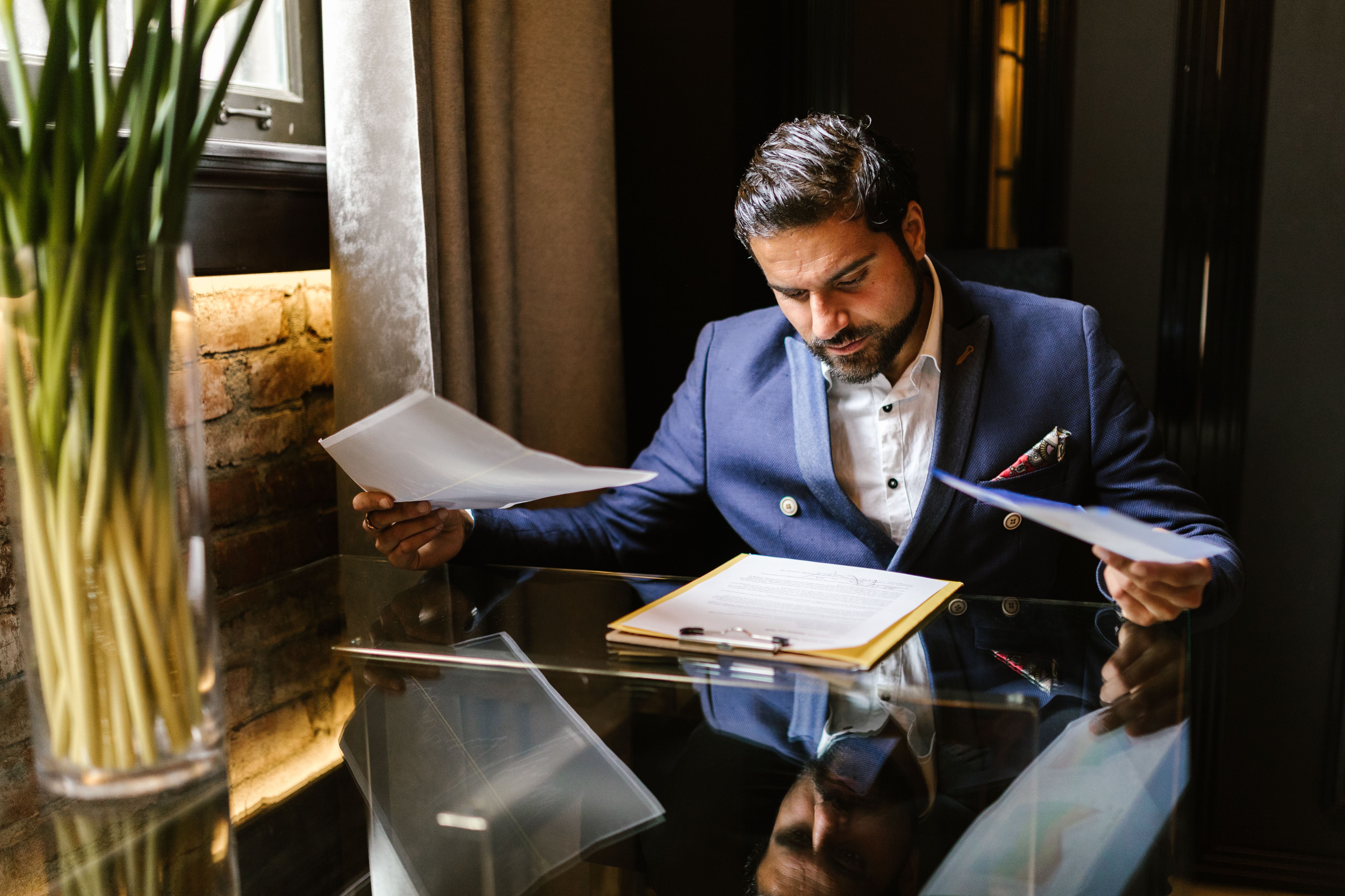 Man in a suit jacket reviewing documents on a glass desk with a clipboard and paper files.