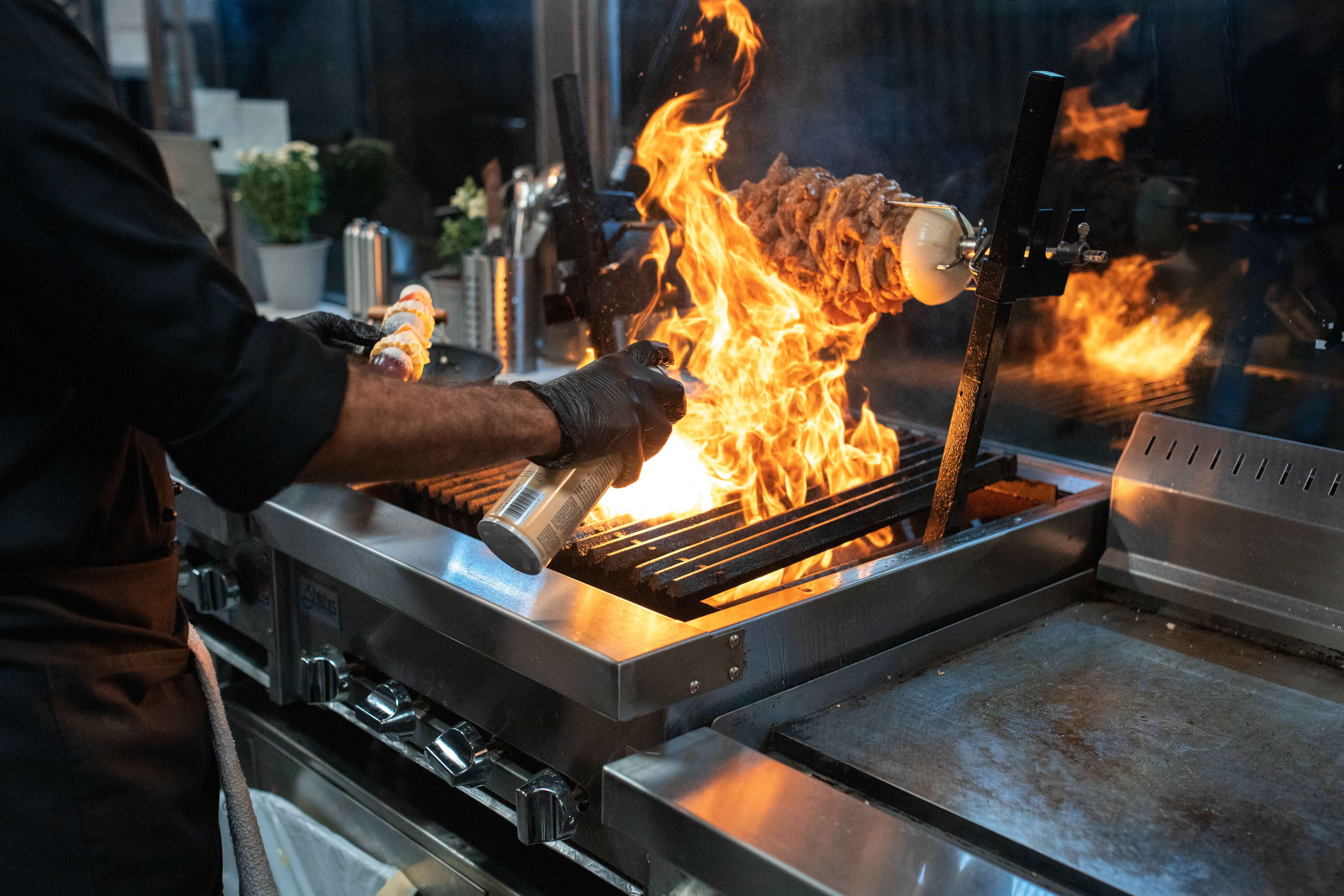 Chef in black gloves spraying oil onto a commercial open-flame grill with meat cooking on a rotisserie.