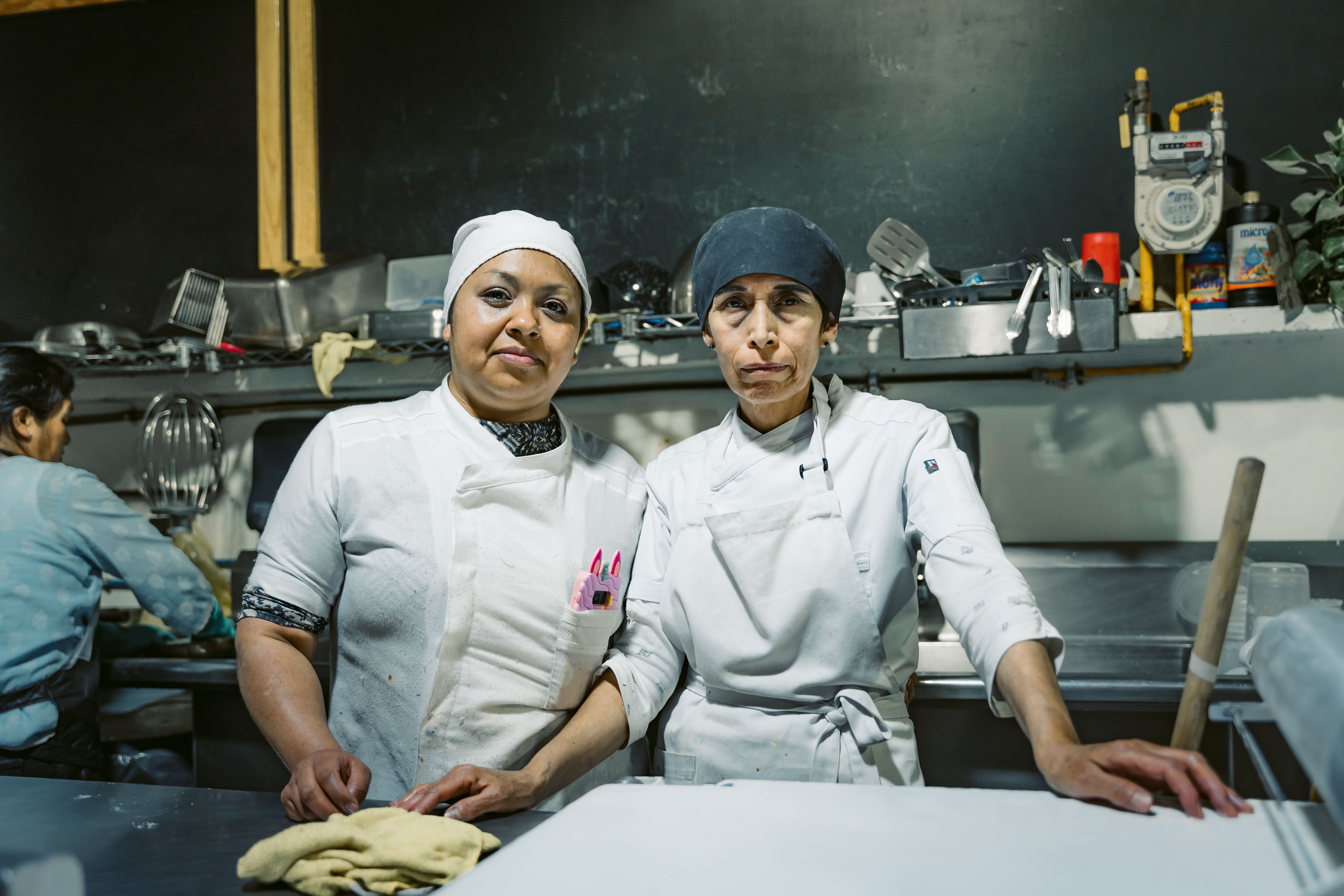 Two female kitchen workers standing together behind a stainless steel prep counter in a commercial kitchen.