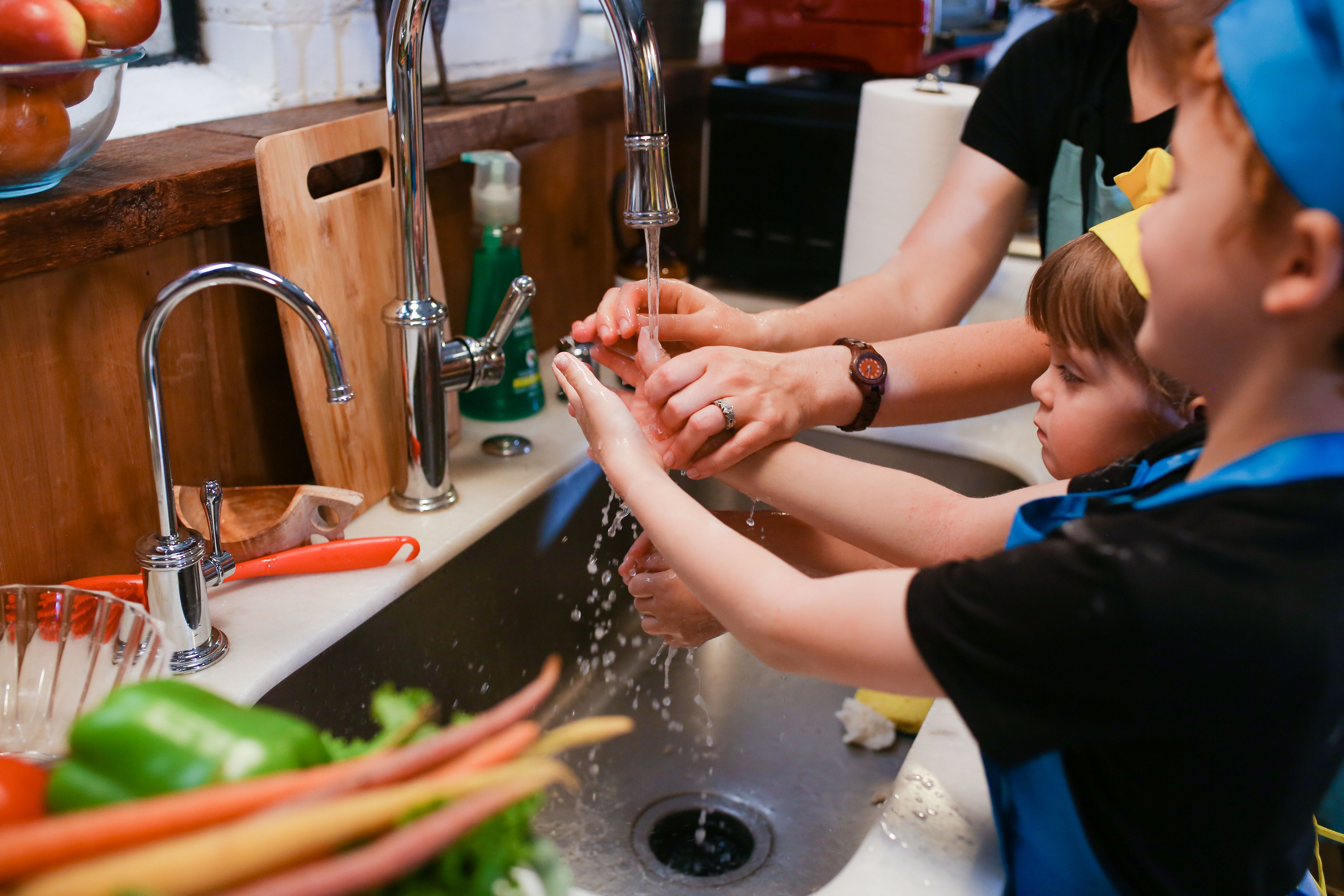 An adult helping two young children wash their hands under running water at a kitchen sink.