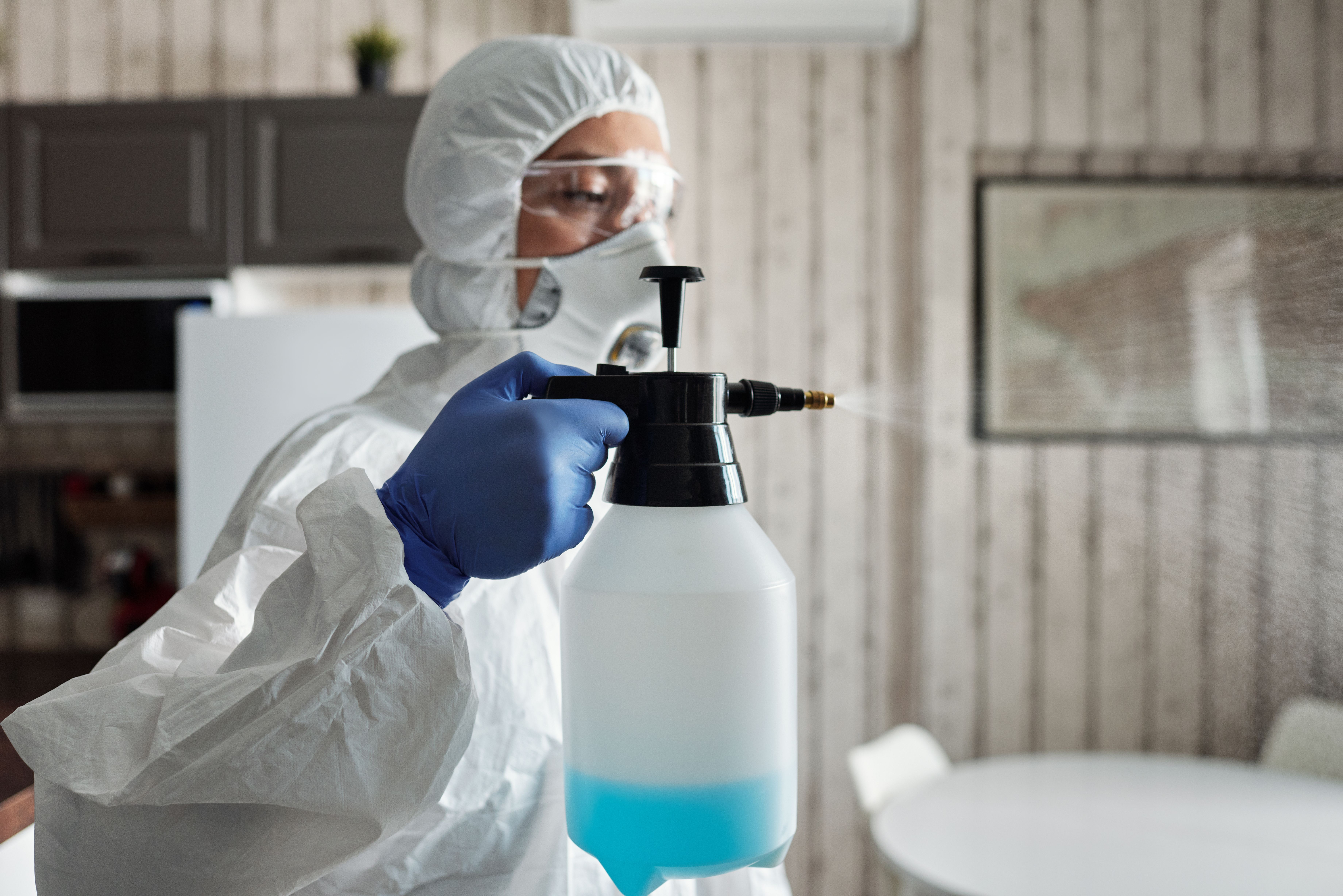 A person in a full white protective hazard suit, face mask, and safety goggles spraying liquid from a pressure canister inside a kitchen.