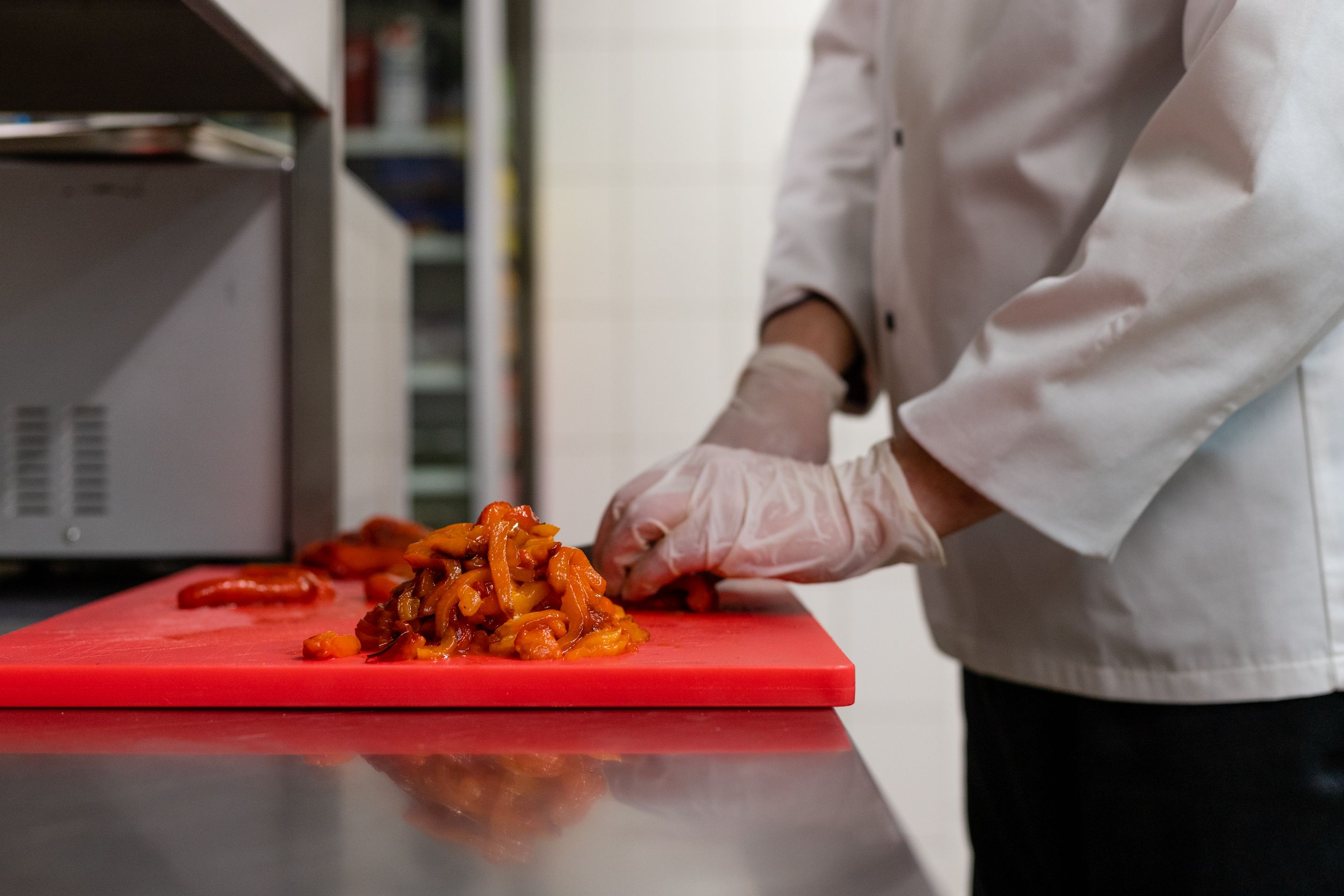 Close-up of a chef wearing disposable gloves and a white uniform slicing red peppers on a bright red cutting board.