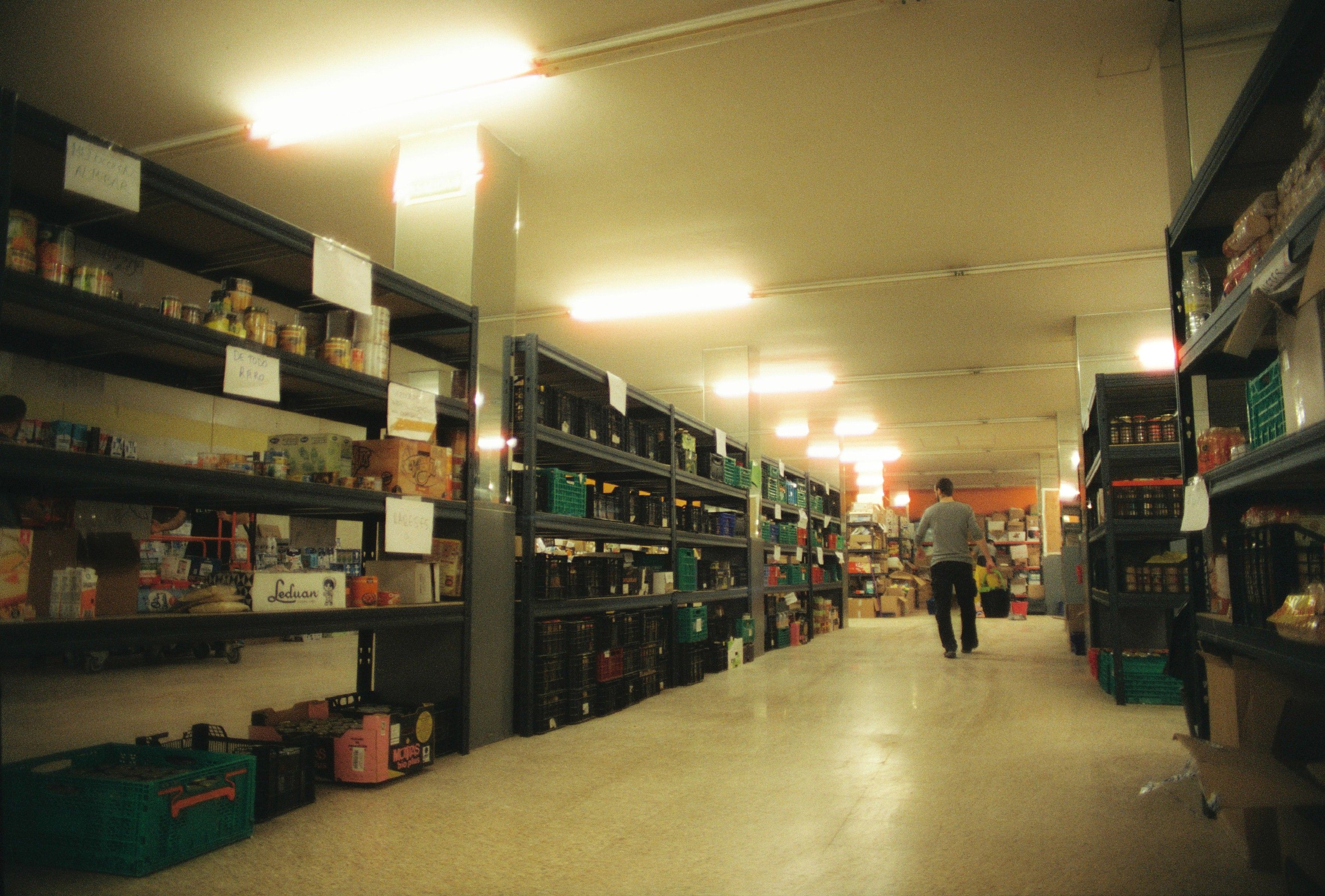 A wide view of a spacious storage room filled with tall metal shelving units holding organized food inventory and boxes, with a person walking down the central aisle.