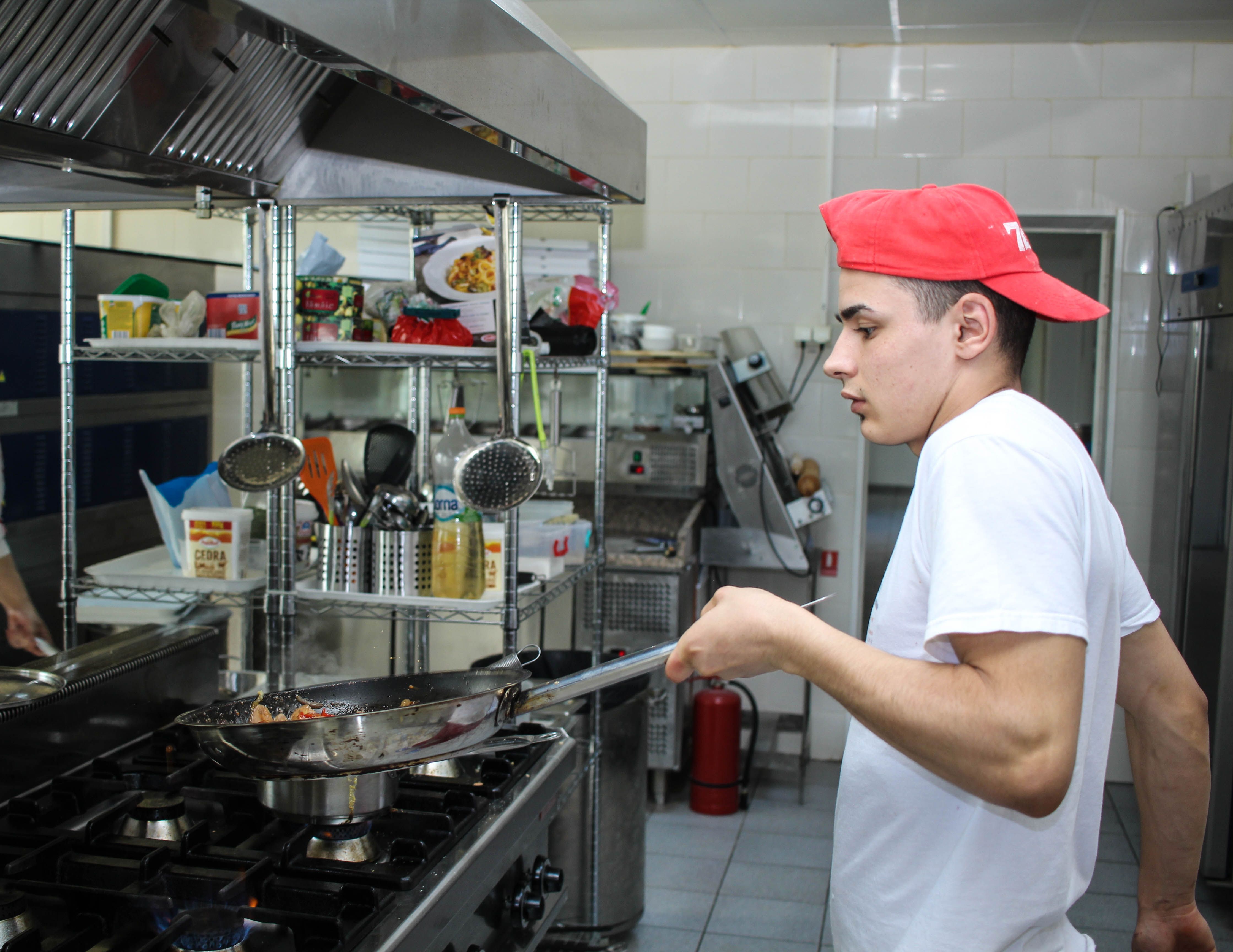 A chef wearing a red cap and white t-shirt tossing ingredients in a frying pan over a commercial gas stove in a kitchen.