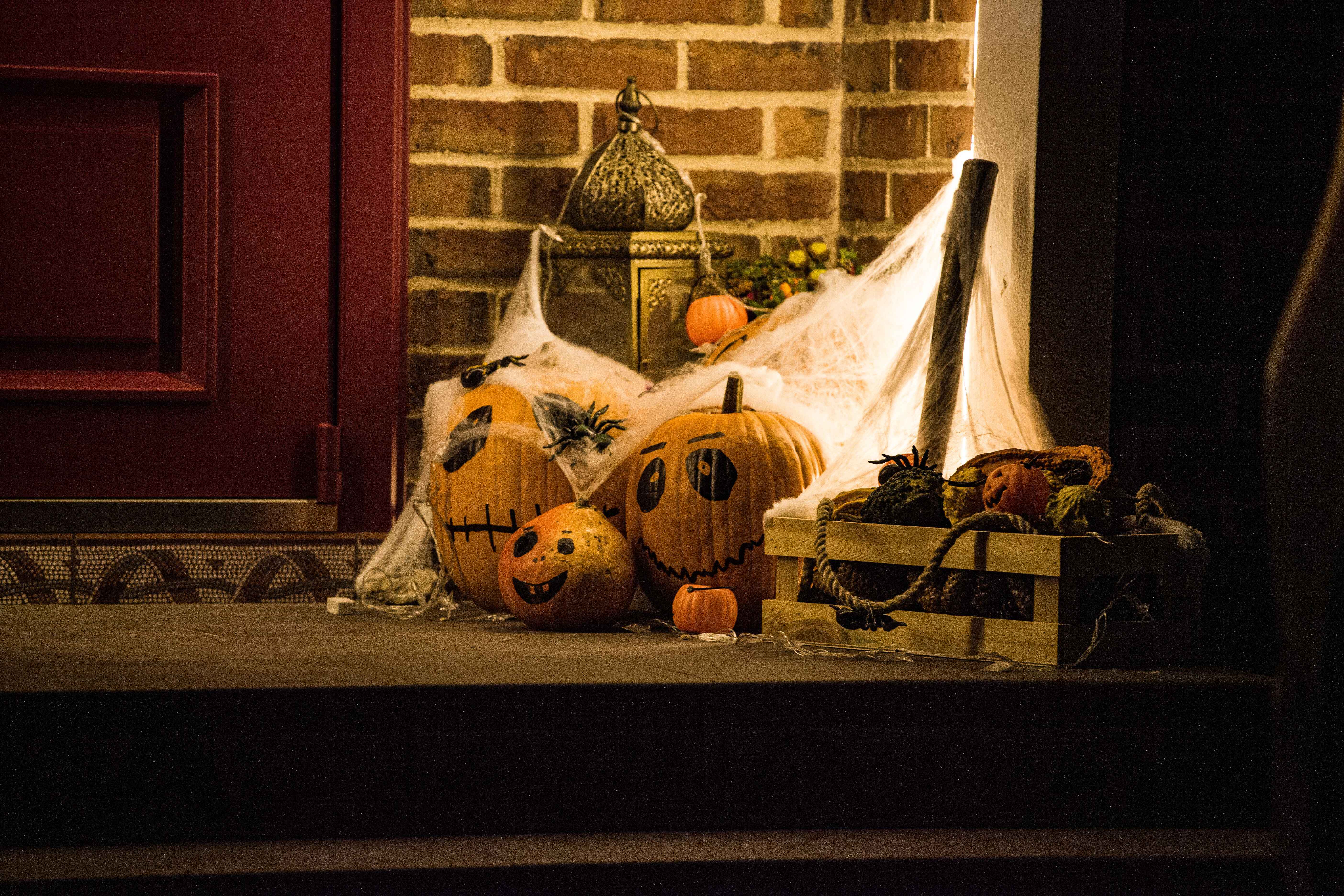 Decorative carved pumpkins with drawn-on faces arranged on a wooden porch doorstep alongside spiderweb decorations and a lantern.