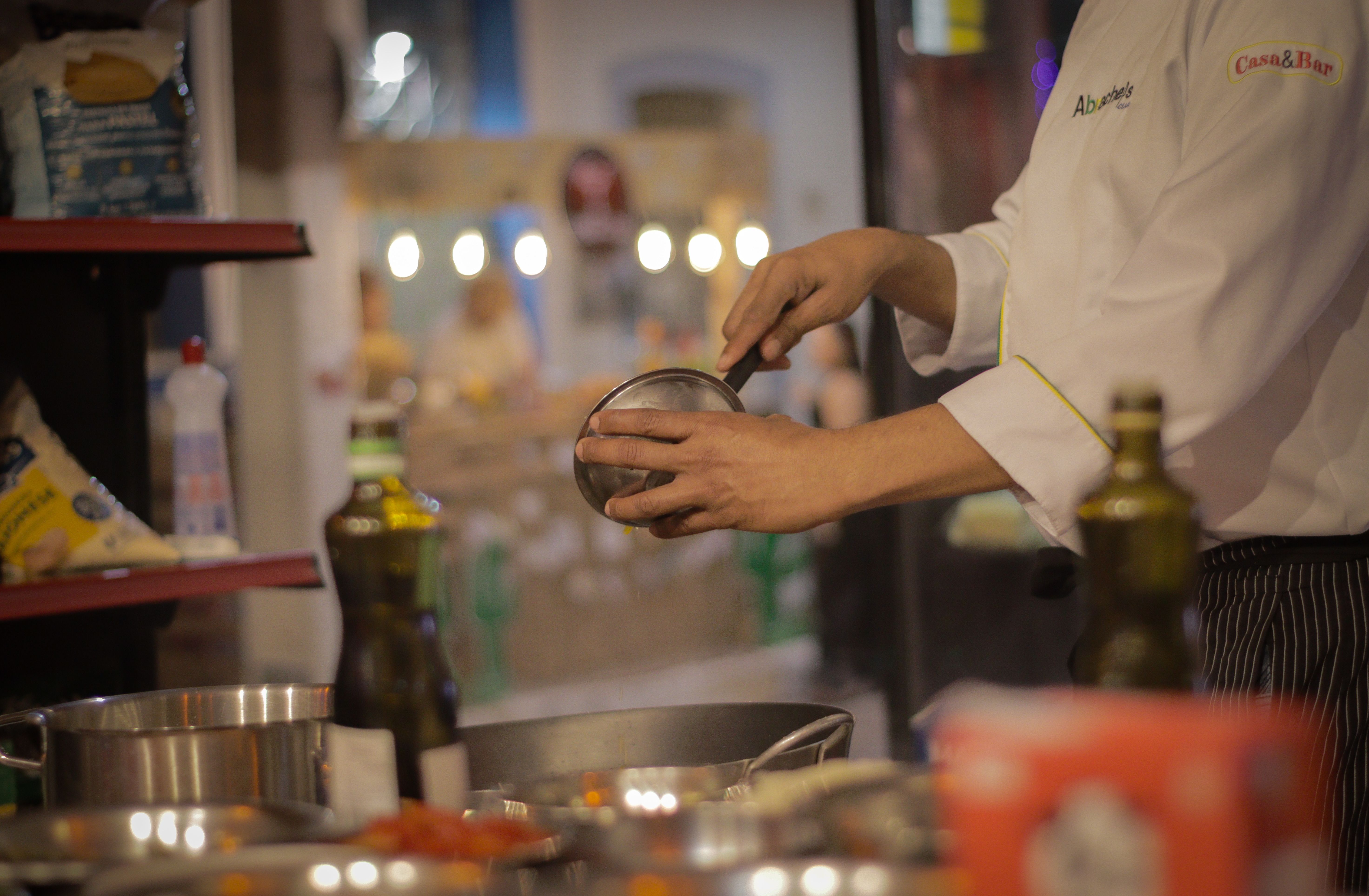 Close-up of a chef in a white uniform using a small bowl to carefully drizzle oil over a dish during preparation in a kitchen.
