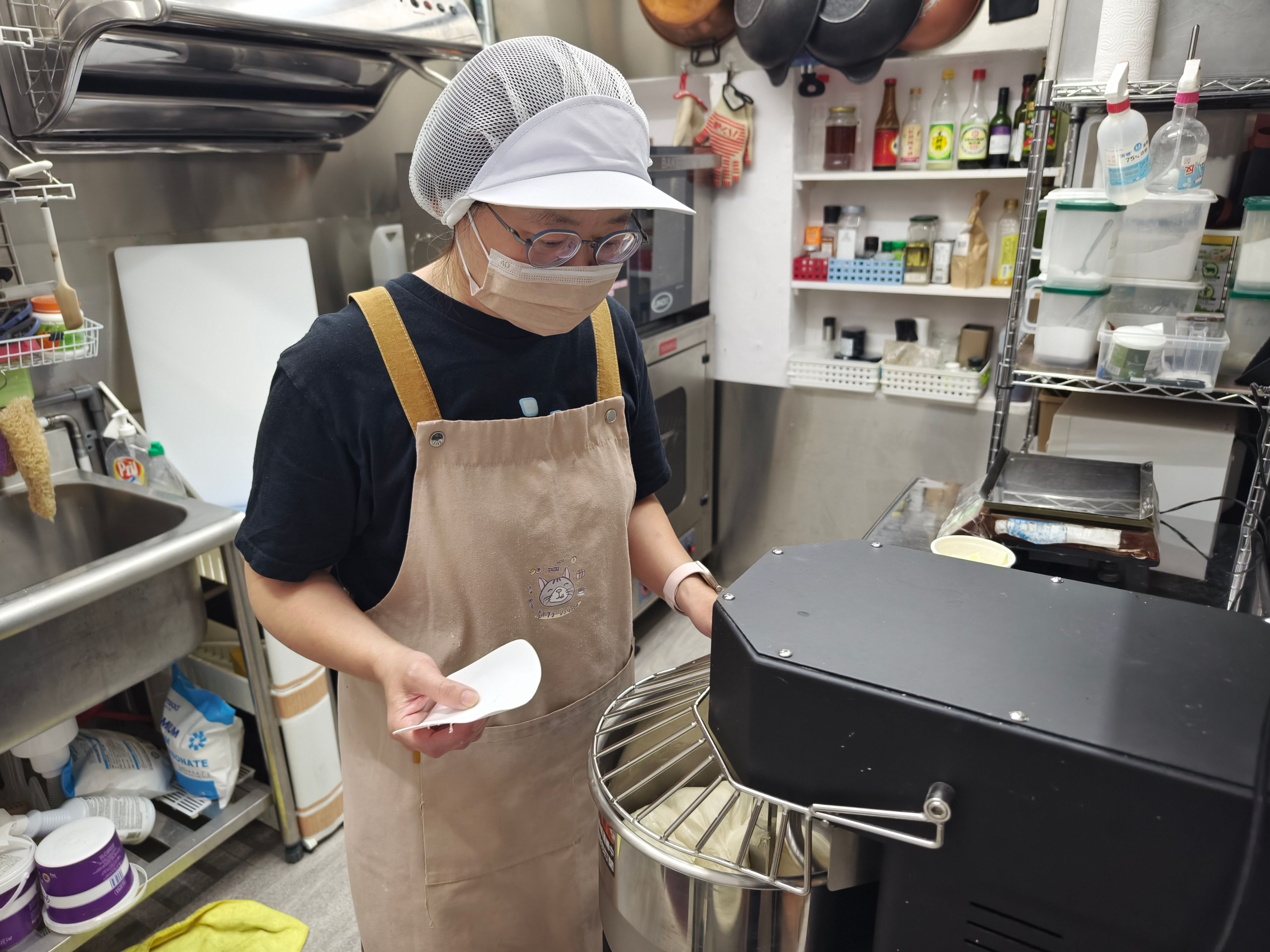 A female baker wearing a hairnet, face mask, and apron standing beside a large commercial dough mixer in a kitchen.