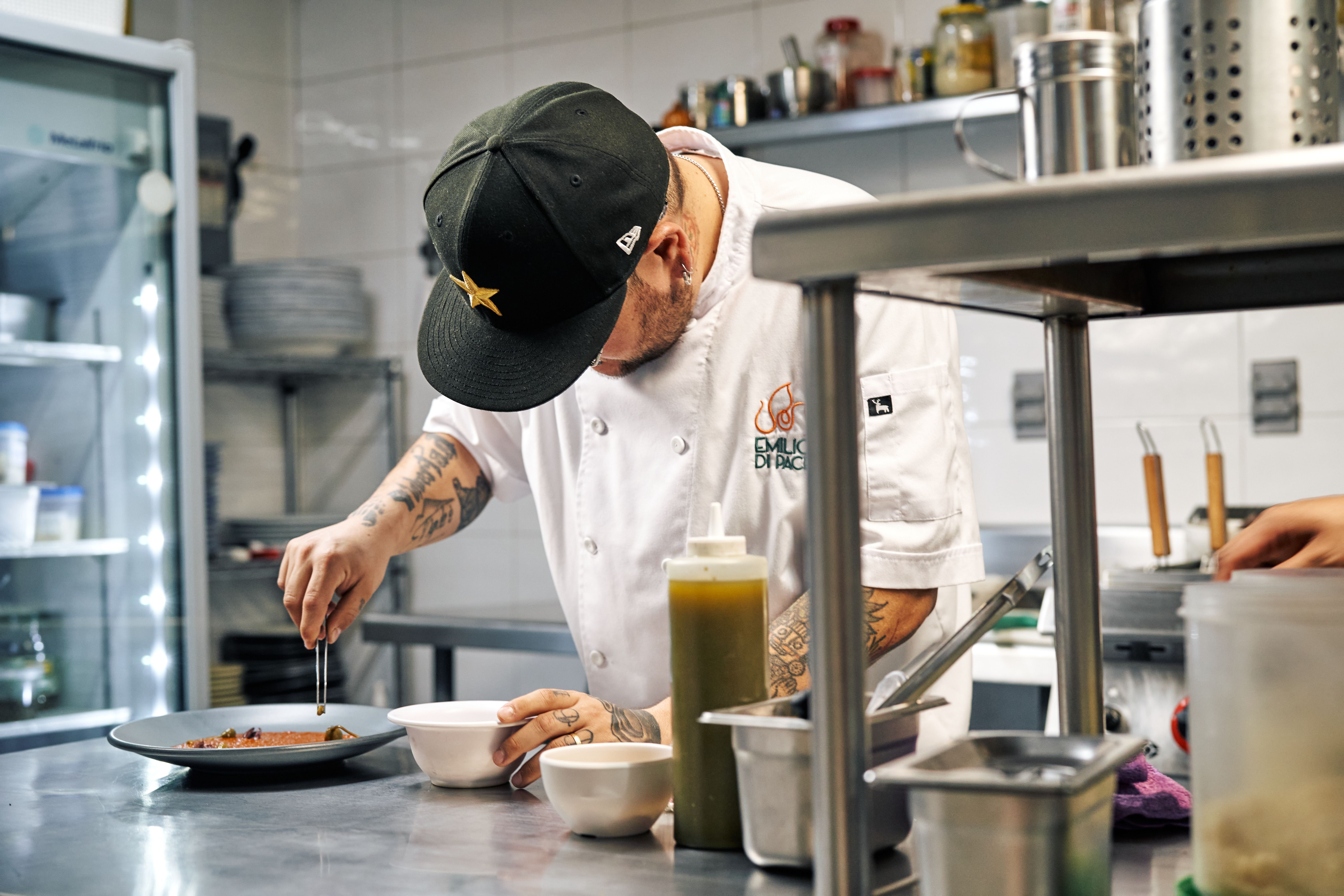 A tattooed chef wearing a black cap and white jacket using tweezers to meticulously plate a dish in a commercial kitchen setting.