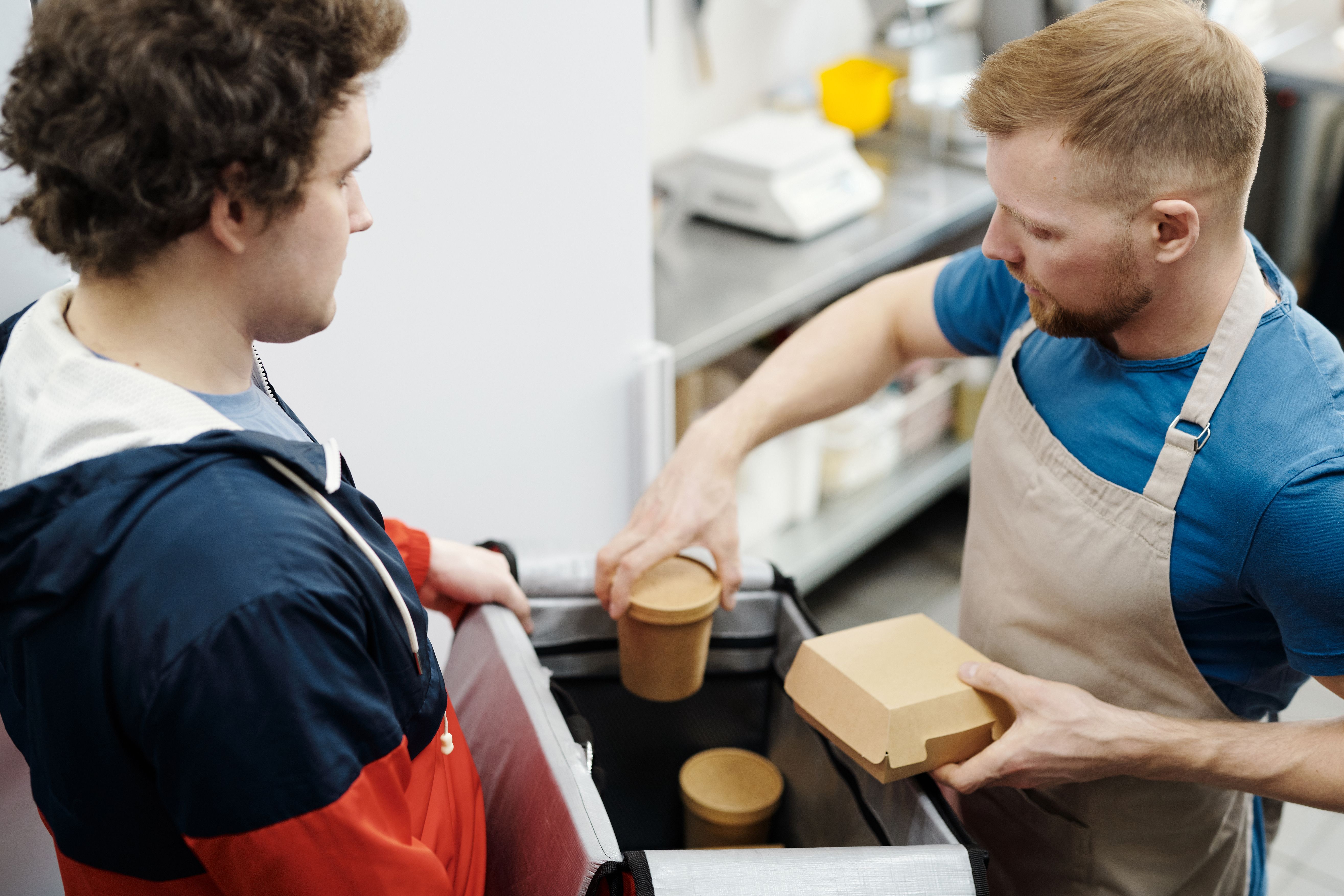 A delivery driver in a jacket holding open an insulated food transport bag while a kitchen staff member wearing an apron places takeout containers inside.
