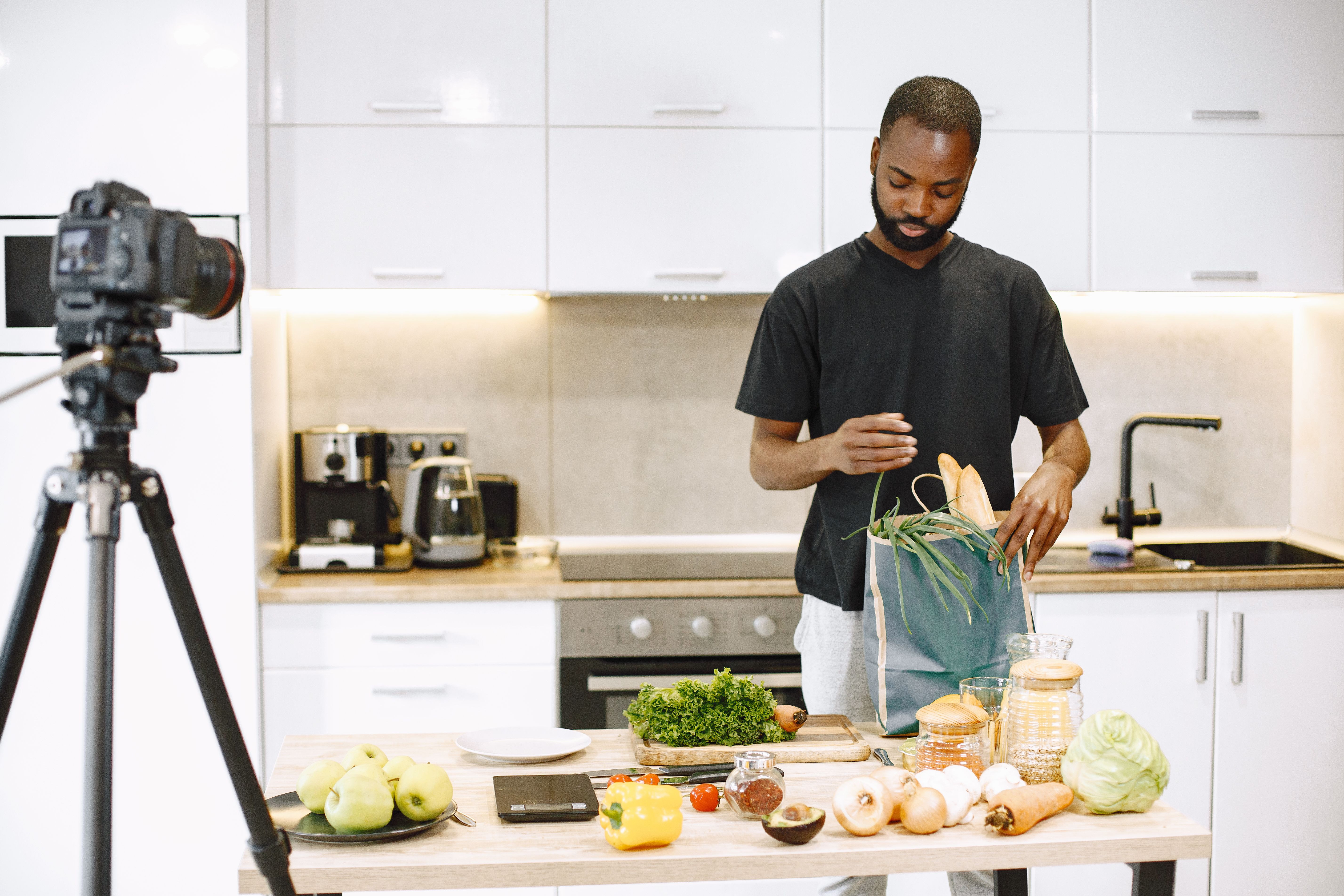 A man standing at a kitchen counter unpacking fresh groceries from a paper bag in front of a camera mounted on a tripod.