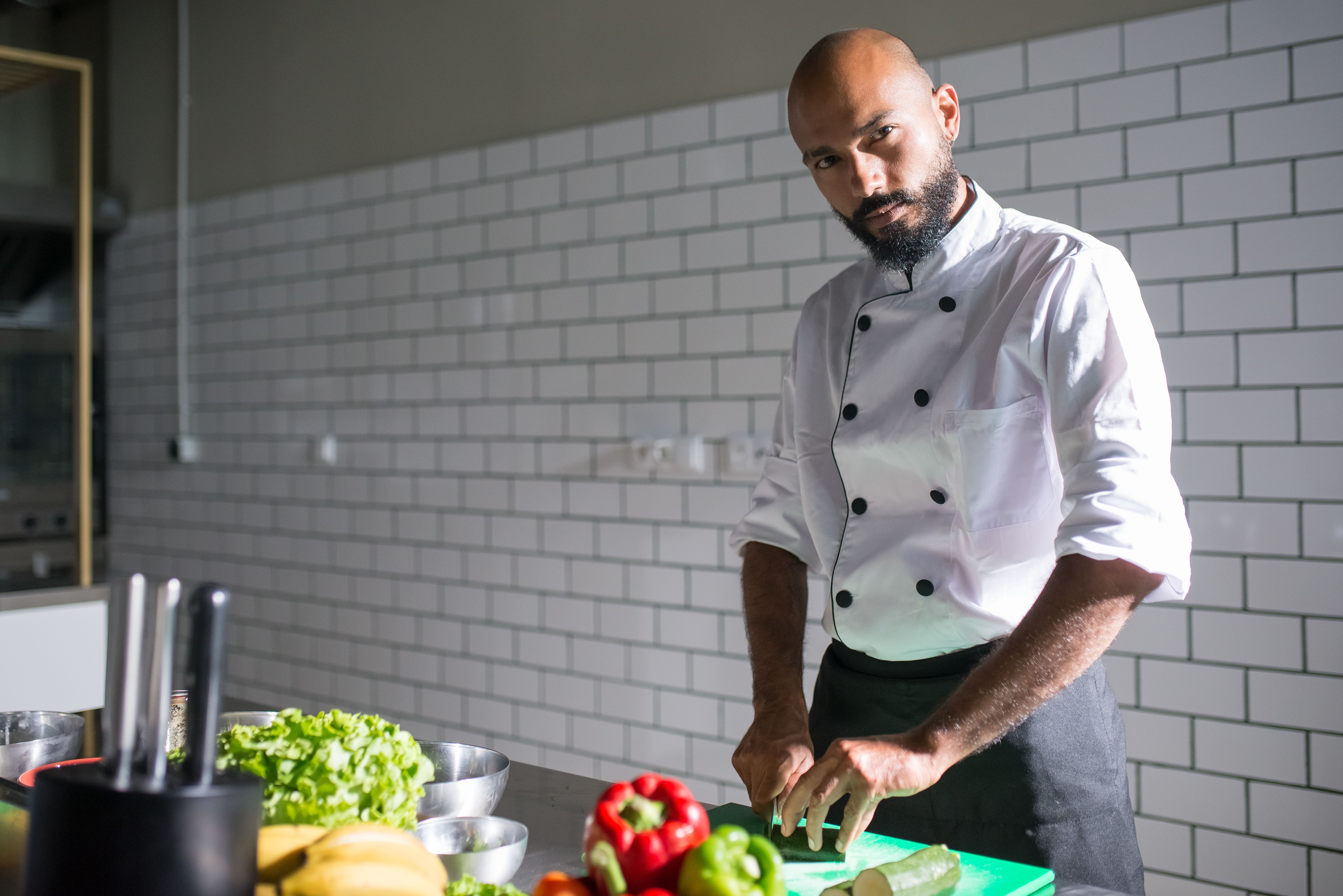 A male chef with a beard wearing a white coat standing in a commercial kitchen with white subway tile, cutting fresh vegetables at a prep counter.