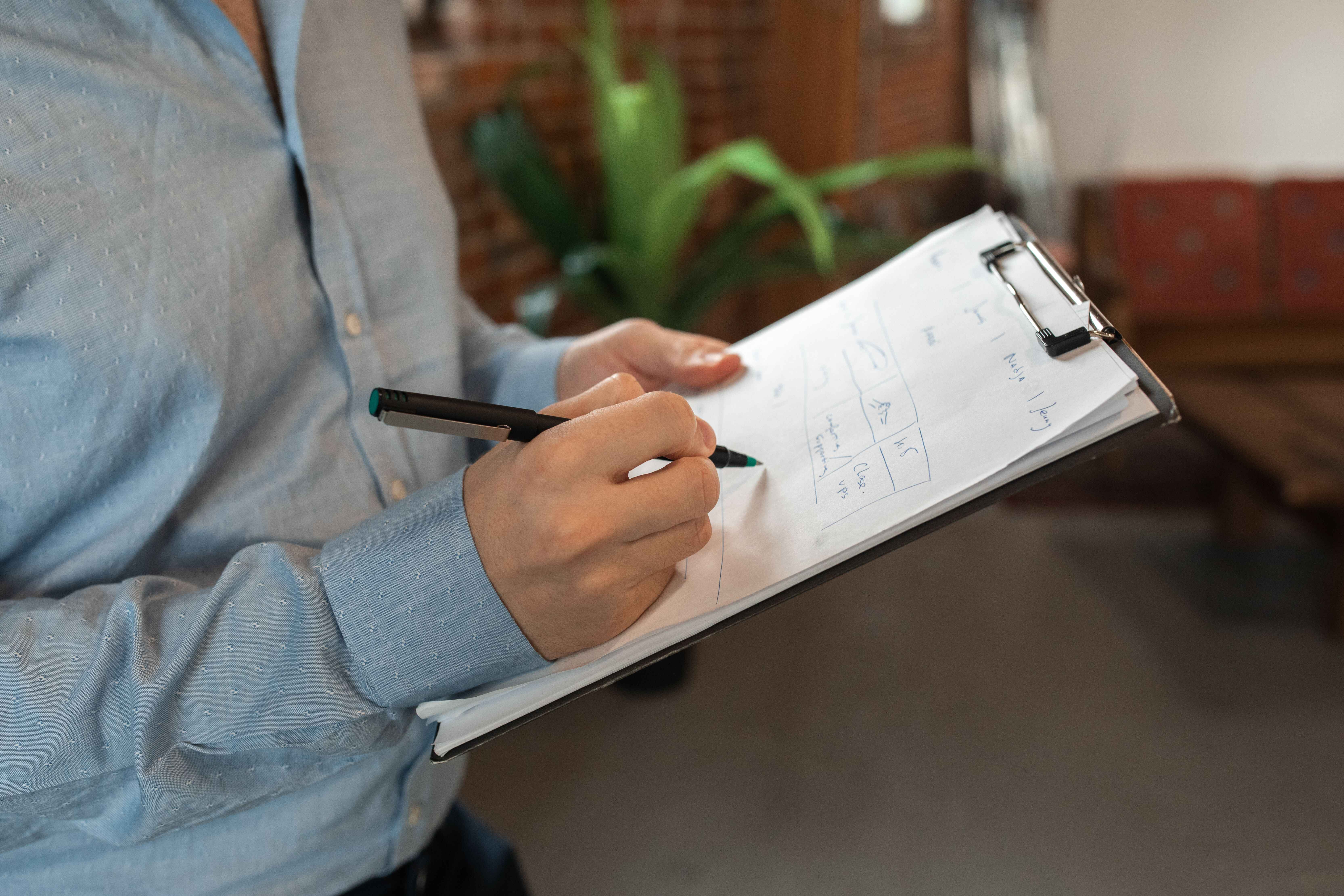 Close-up of a person in a blue button-down shirt holding a clipboard and using a black pen to fill out a printed audit checklist.