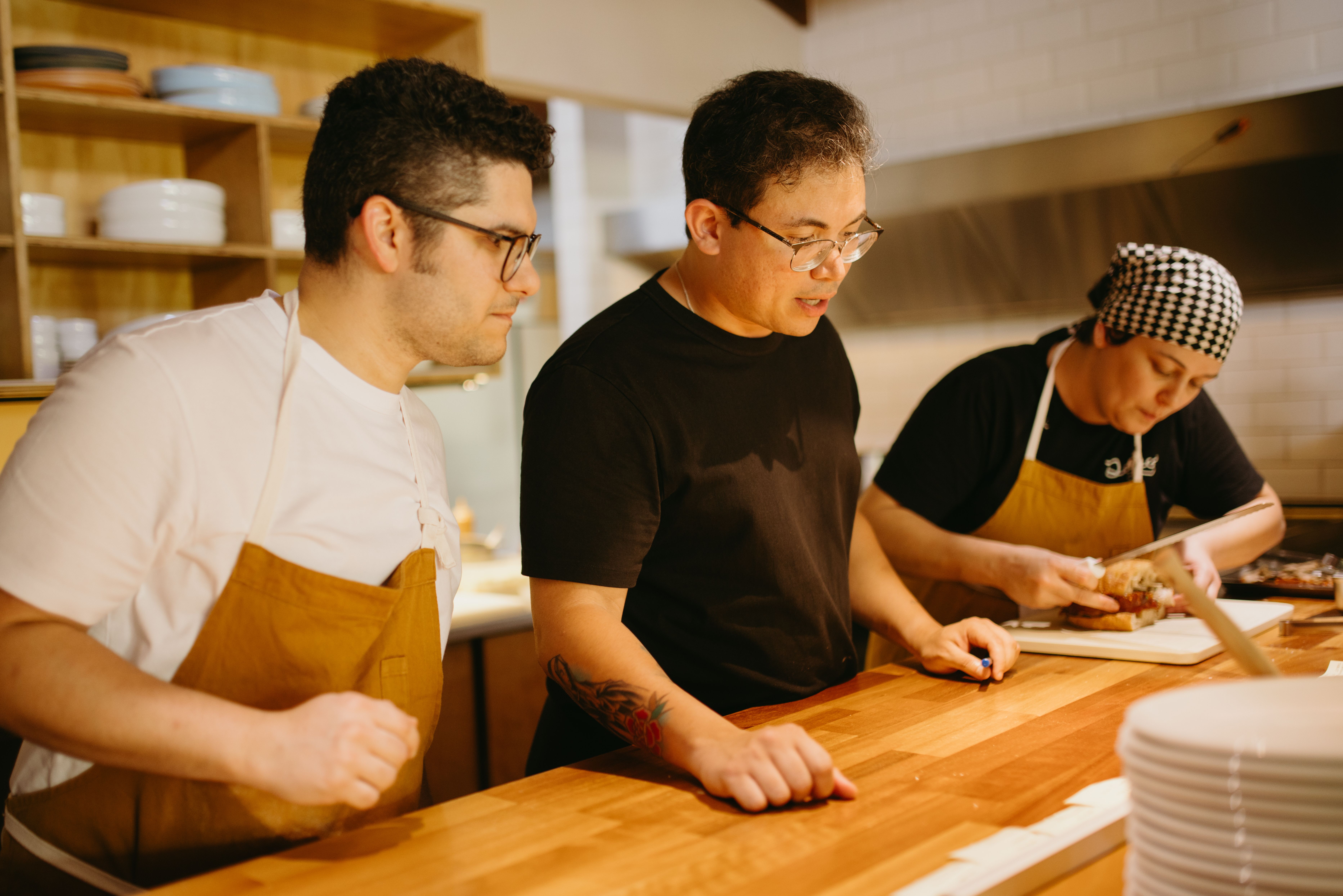 A instructor wearing a black shirt and glasses standing in a commercial kitchen next to a student, demonstrating food preparation techniques while a colleague works in the background.