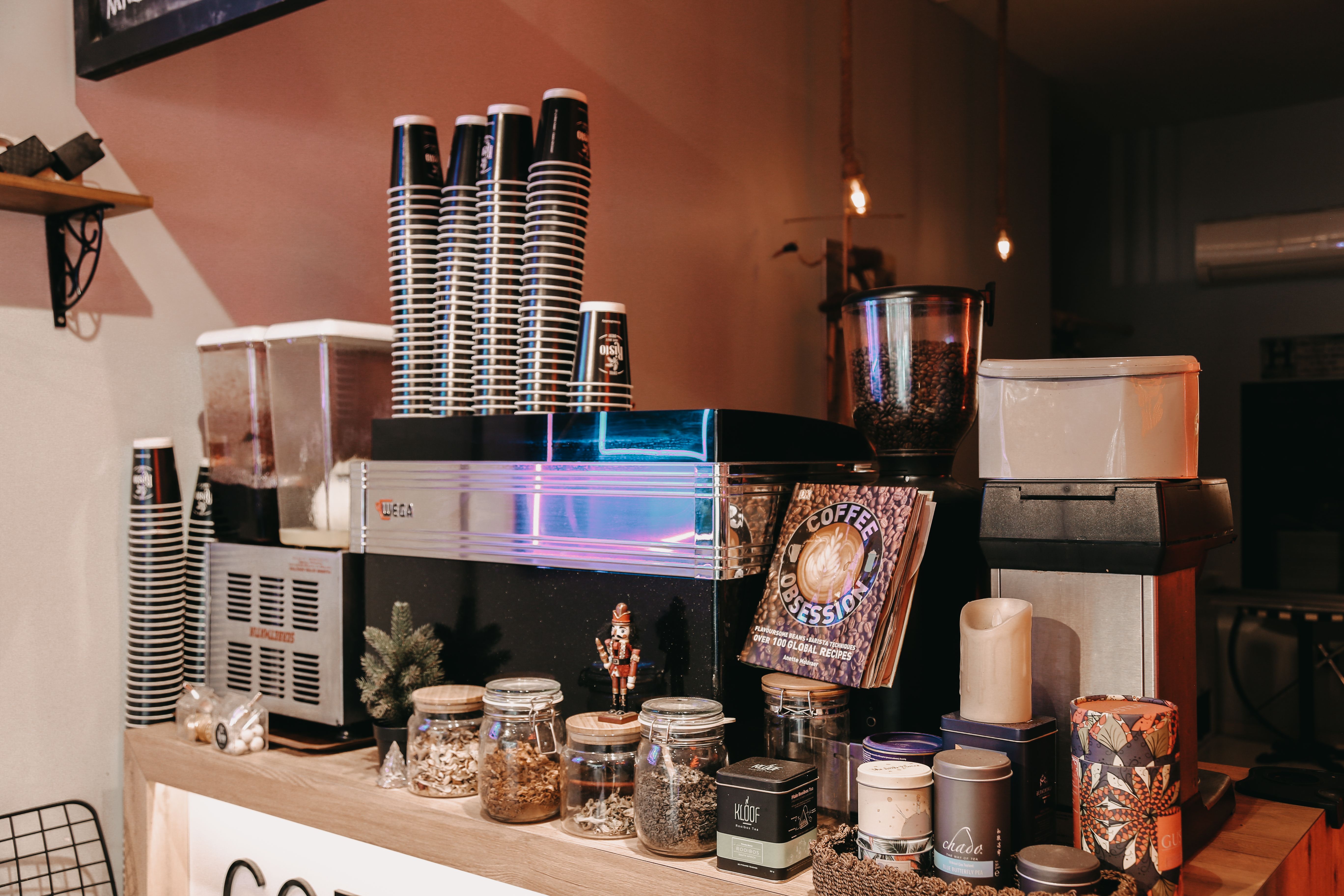 An espresso machine with stacks of takeaway cups on top, set up on a café counter alongside jars of tea and coffee accessories.