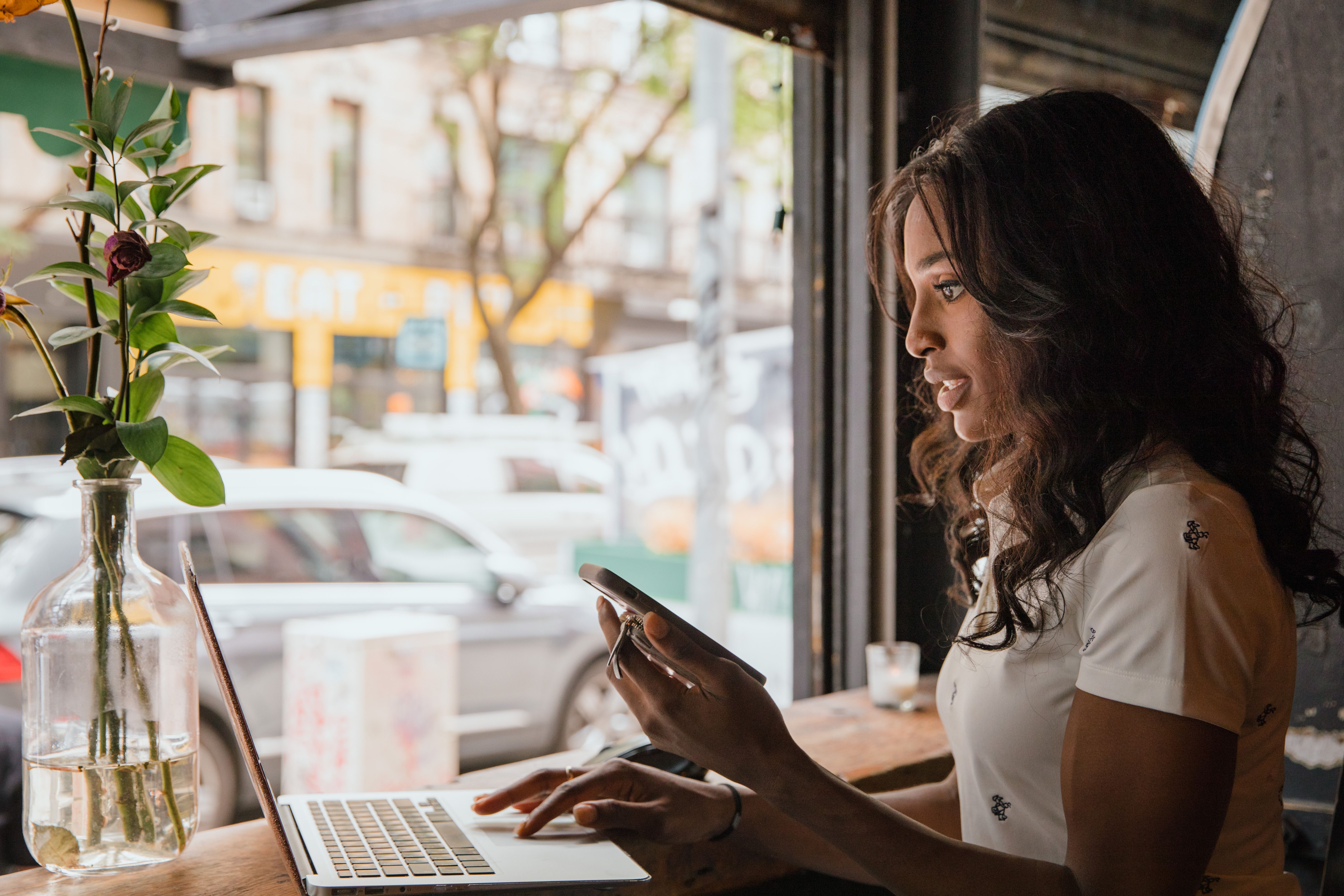 A woman sitting at a counter near a window, typing on a laptop while holding a smartphone in her hand.