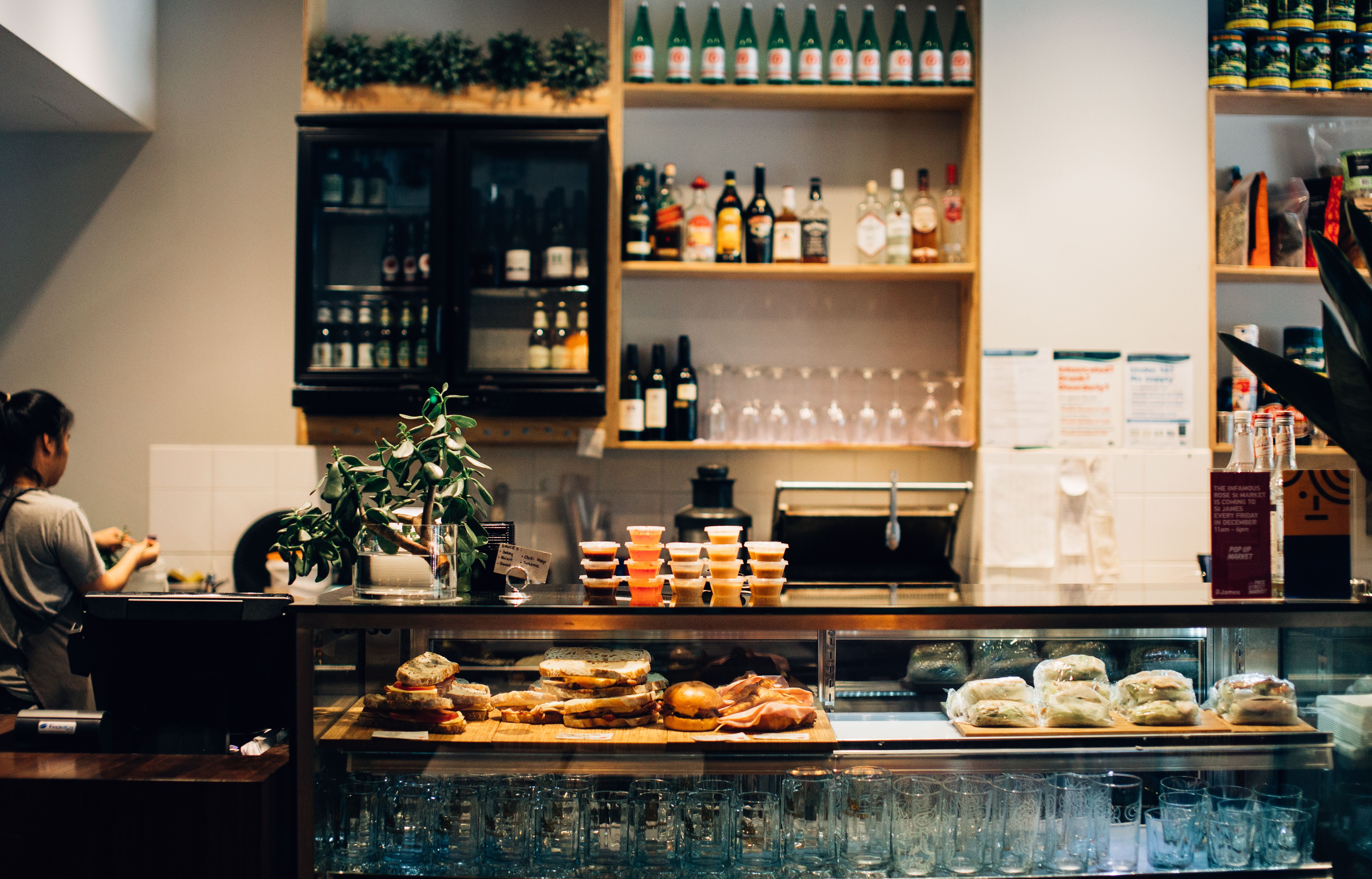 A café counter display case featuring fresh sandwiches and small plastic condiment cups, with alcohol bottles and glassware arranged on shelves behind it.