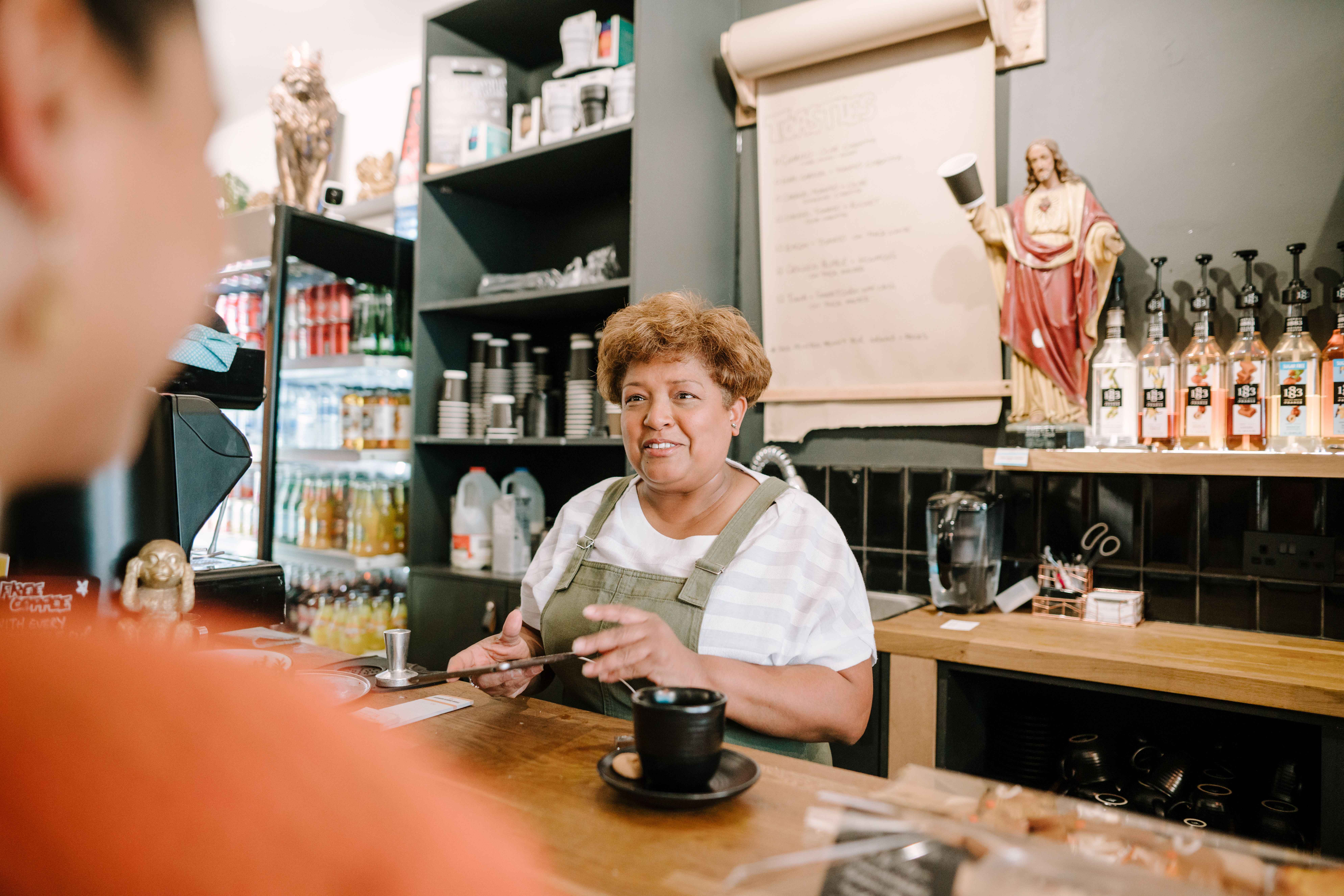 A female cafe worker in an apron behind a wooden counter speaking to a customer, with syrup bottles and a menu displayed behind her.