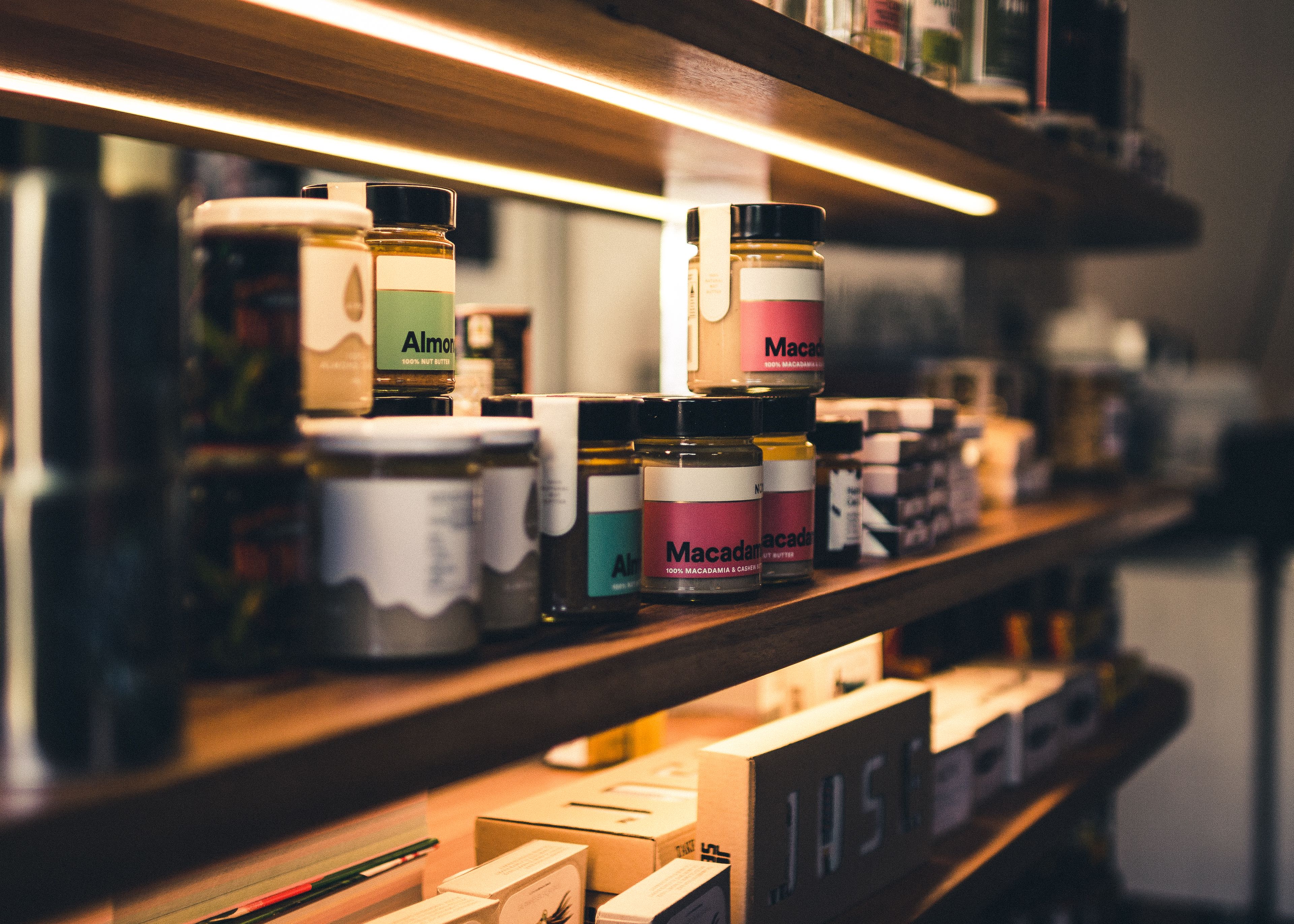 Jars of nut butters labeled Almond and Macadamia arranged on wooden retail store shelves lit by warm strip lighting.