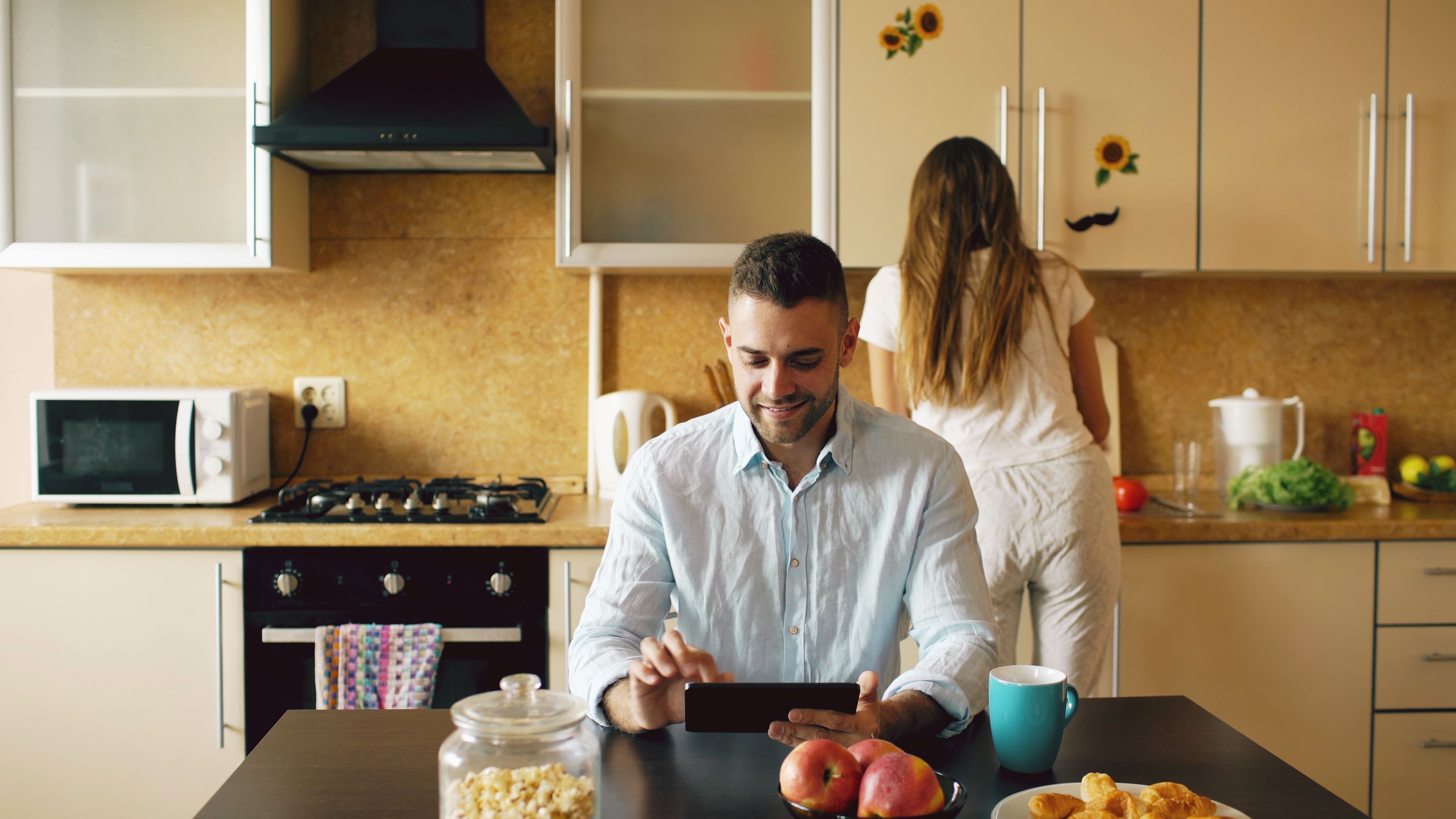 A man sitting at a kitchen table using a smartphone while a woman stands in the background near modern kitchen cabinets.