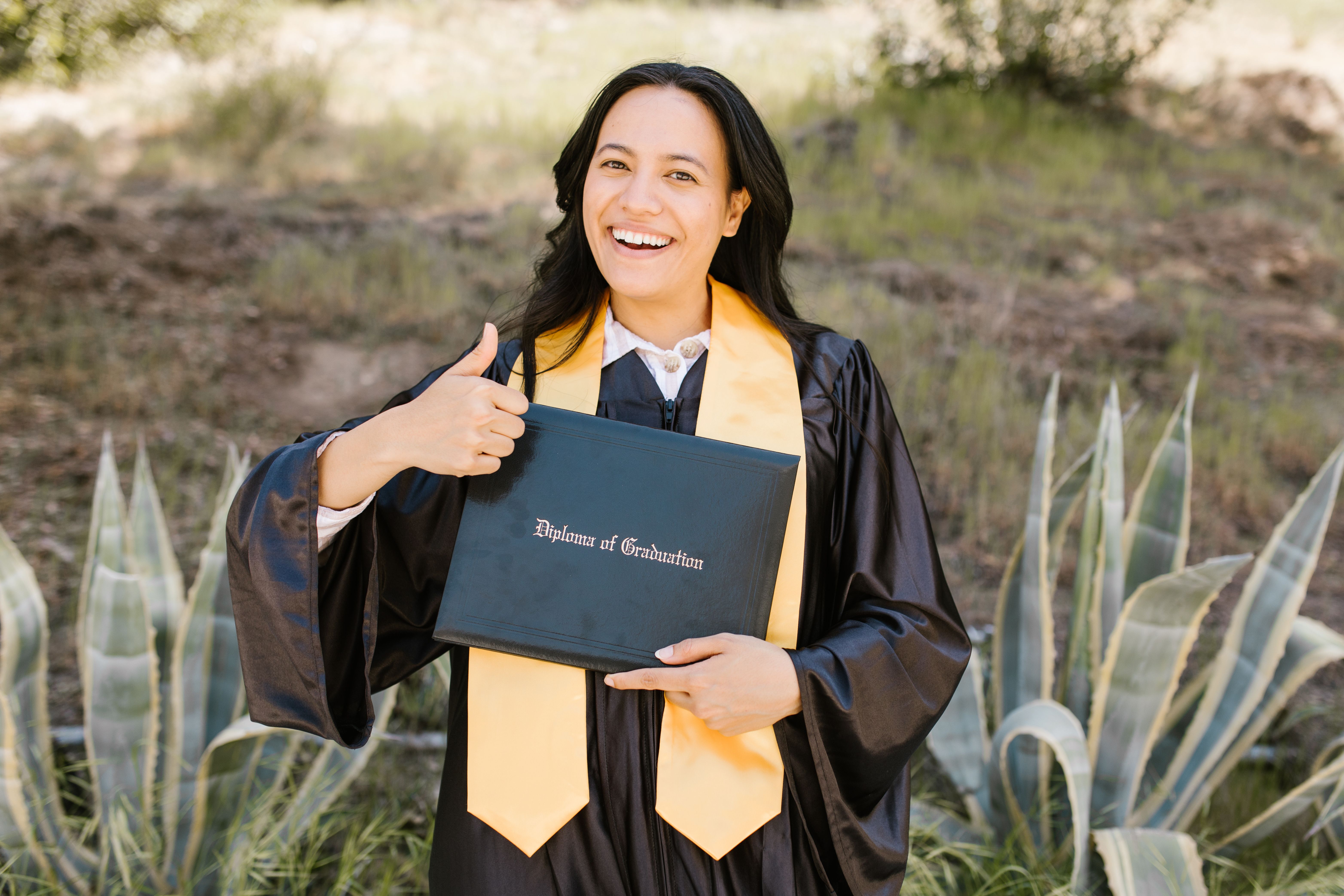 A smiling graduate in a black gown and yellow sash giving a thumbs-up while holding a diploma cover outdoors.