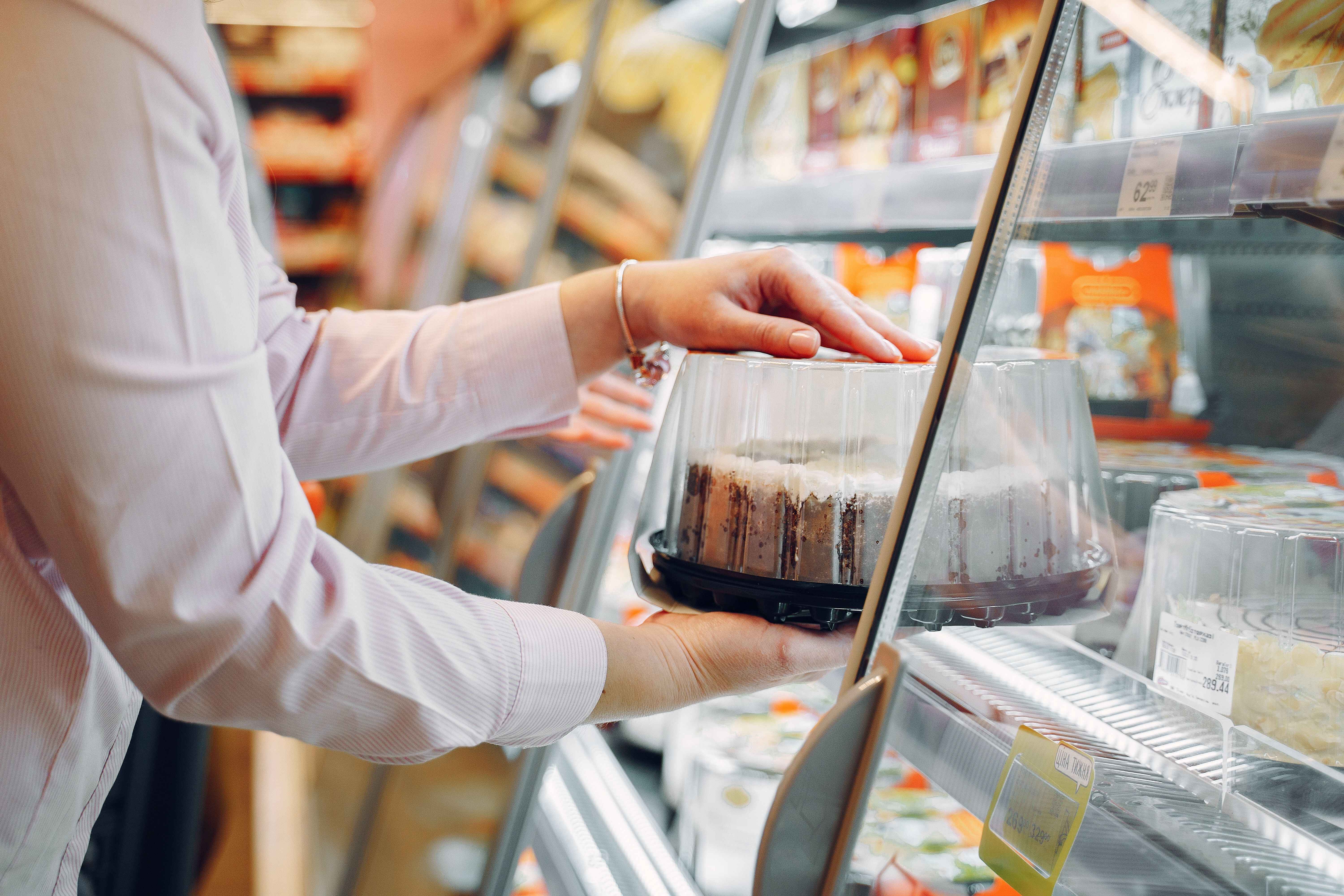 A customer reaching into a refrigerated display case to select a packaged cake in a clear plastic container.