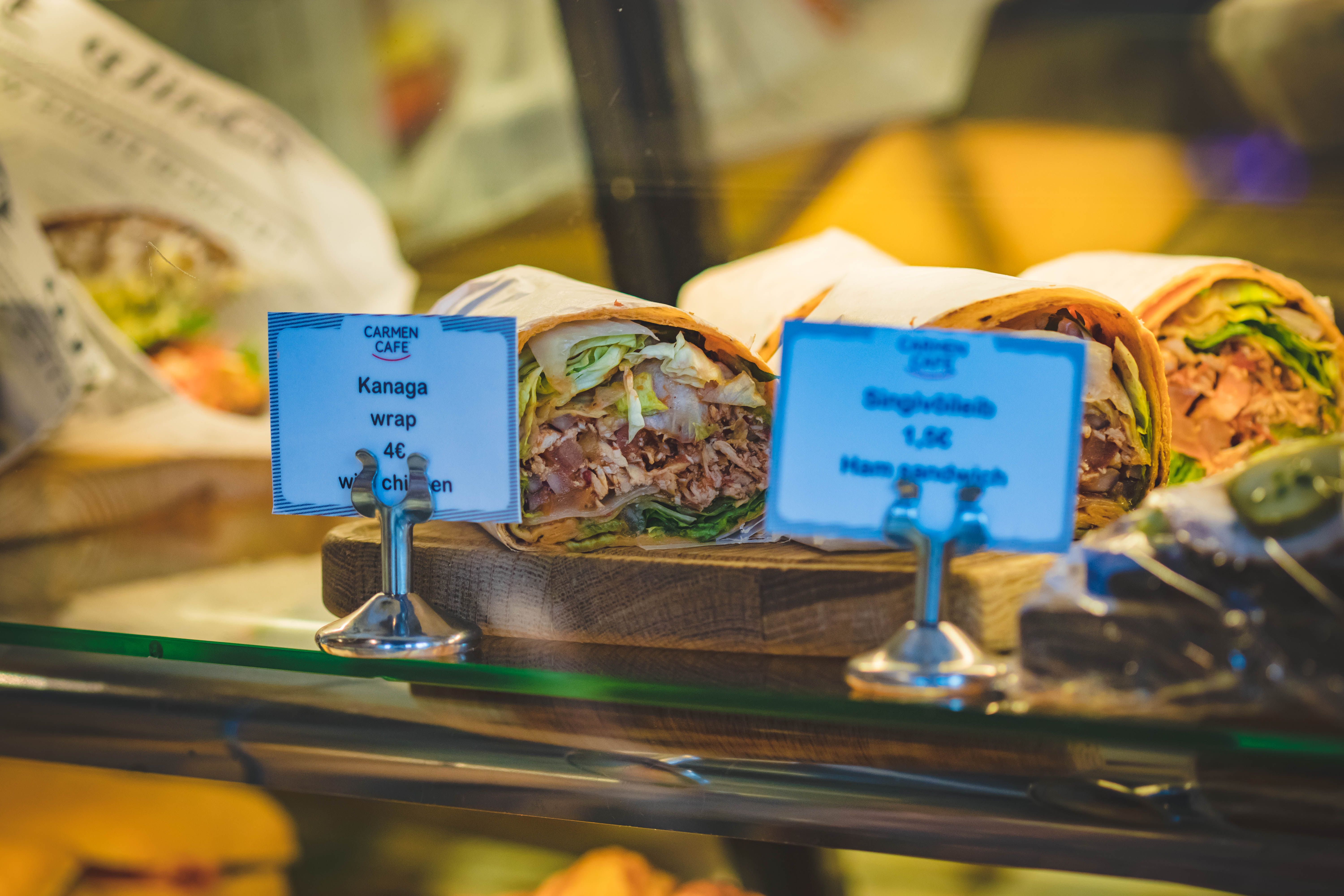 Pre-packaged food wraps and sandwiches on display behind a bakery glass counter with small price signs.