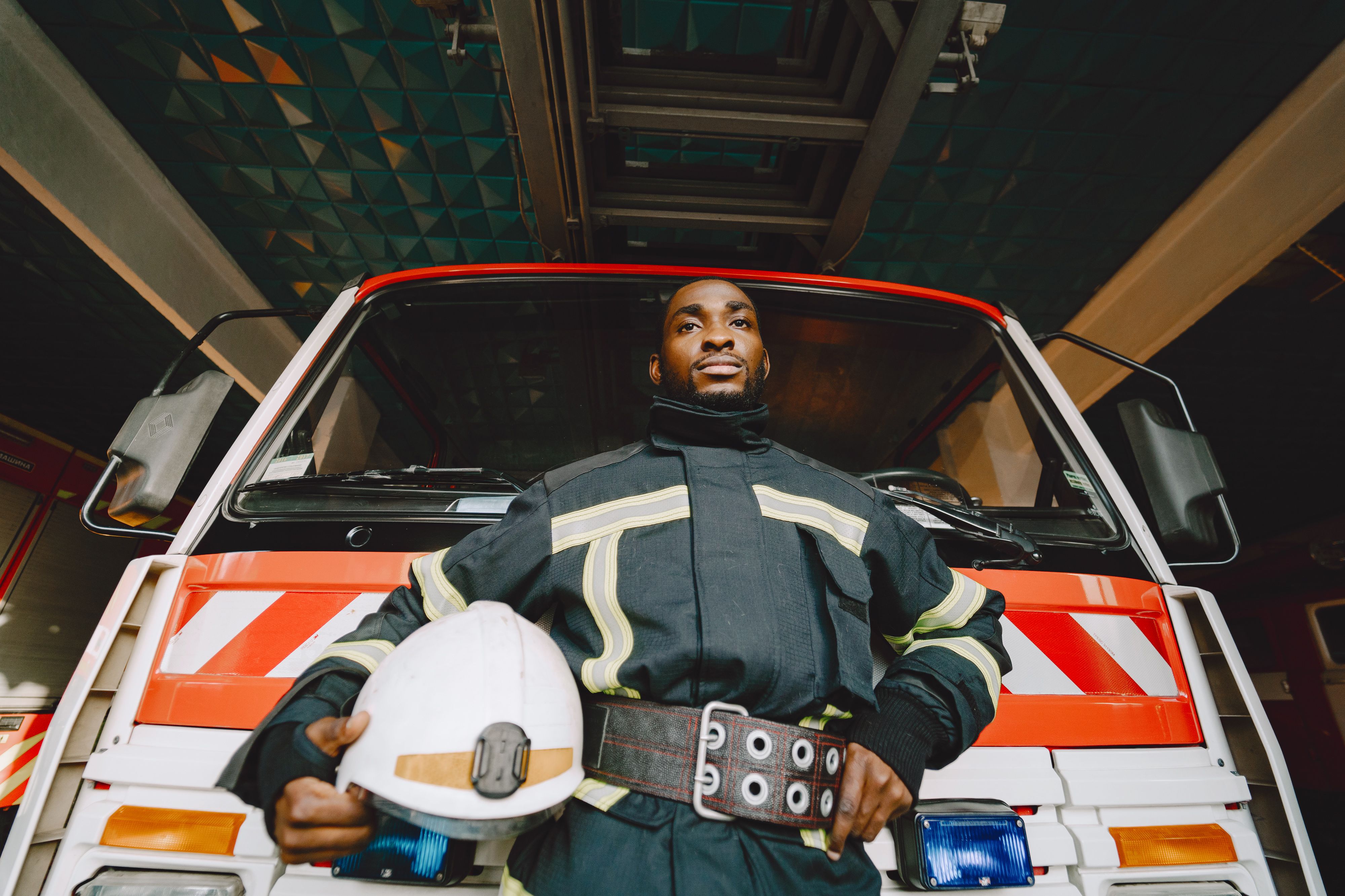 A firefighter in protective gear and wide belt holding a hard hat and standing in front of a red fire truck.