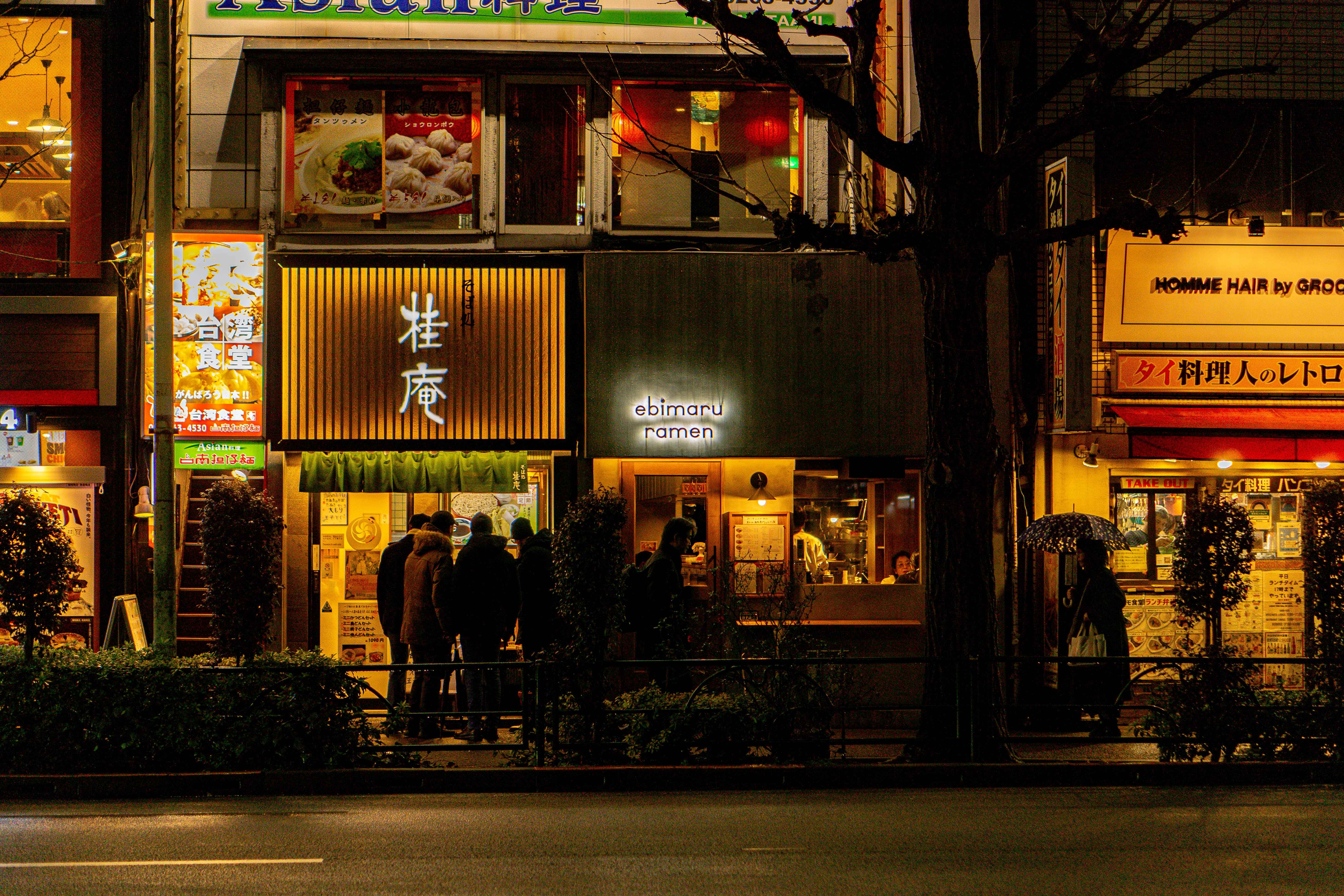Exterior view of illuminated restaurant storefronts and ramen shops along a city street at night.