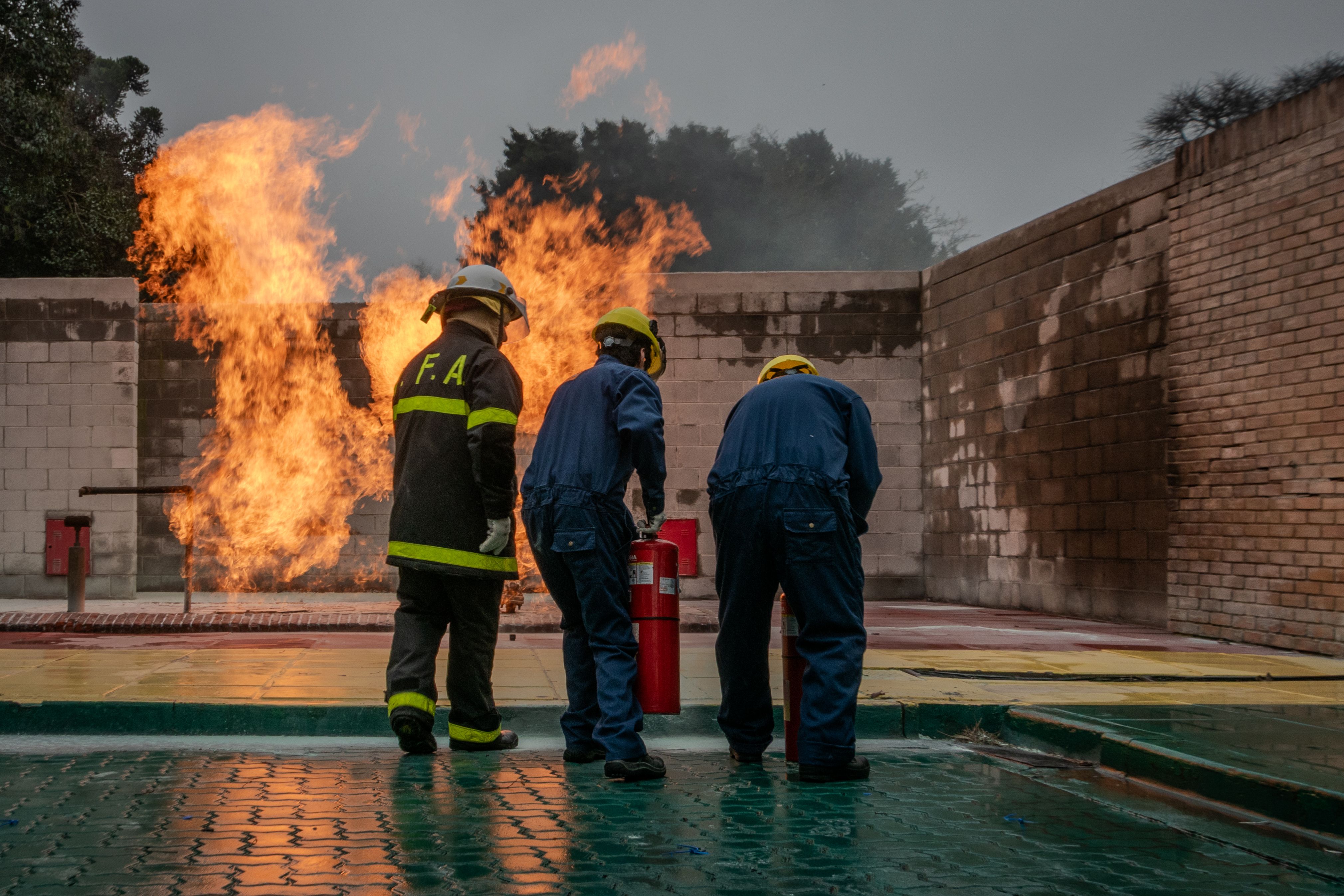 Three firefighters wearing helmets and protective suits conducting a controlled burn exercise with fire extinguishers outdoors.
